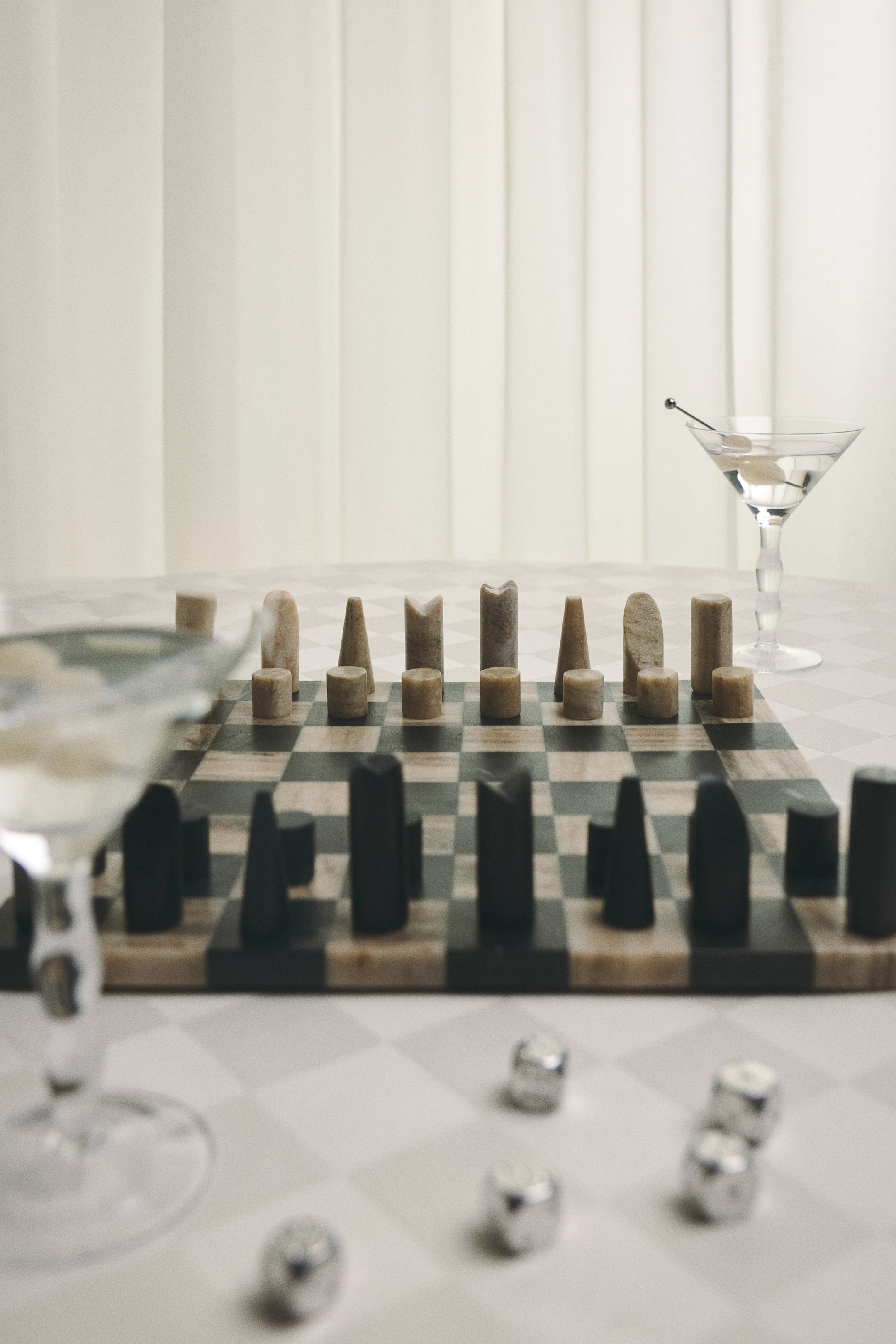 View larger image: A minimalist marble chess set features a square board with alternating light beige and dark charcoal-grey checkered squares. Sculptural, geometric chess pieces carved from natural marble are standing in starting positions, displaying their unique, varied textures in matching light beige and dark grey tones.