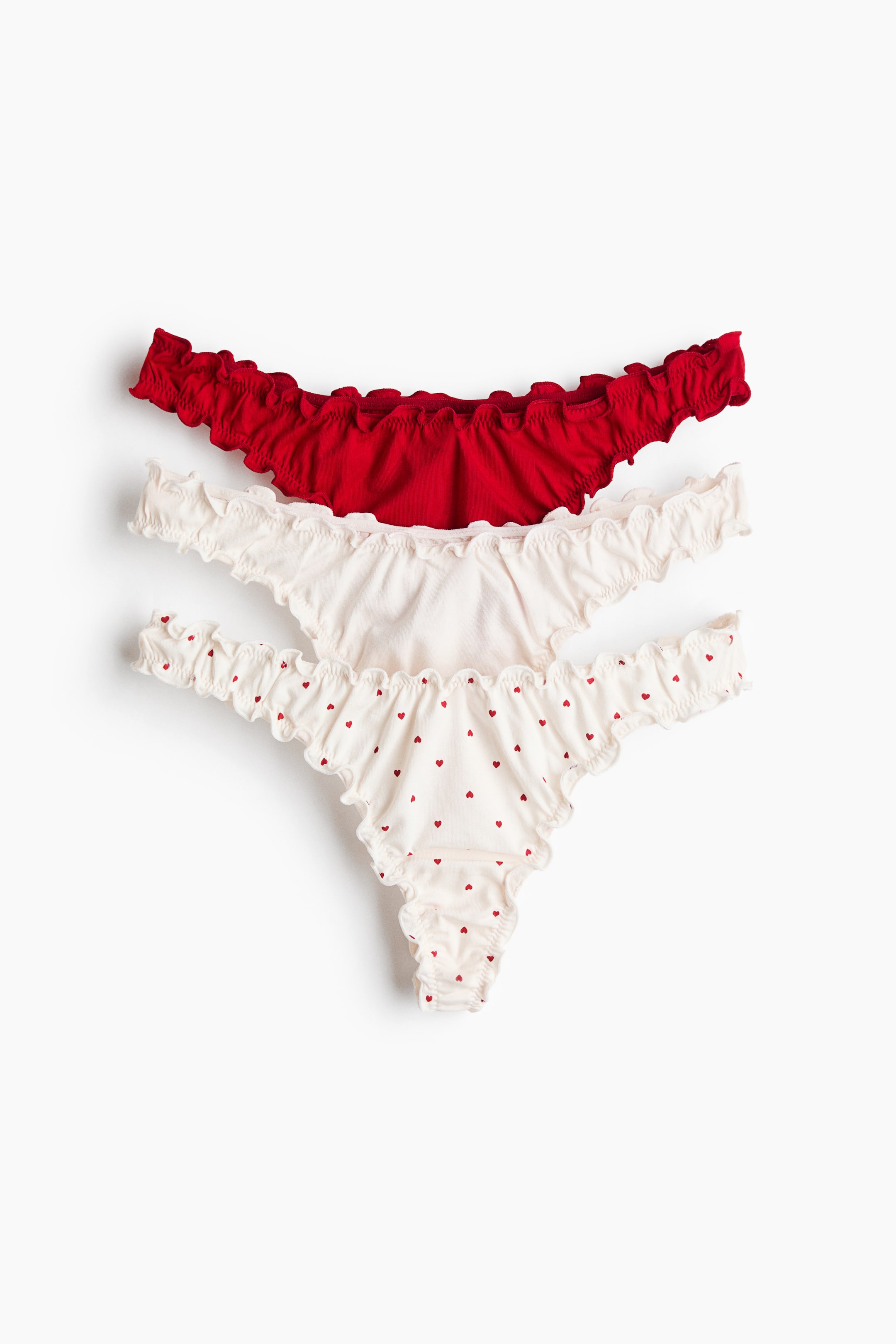 View larger image: Three G-strings are laid flat and shown from the front, featuring a low-rise, V-shaped waist, and ruffled overlocked edges. The set includes one solid red pair, one solid off-white pair, and one off-white pair with a repeating pattern of small red hearts.