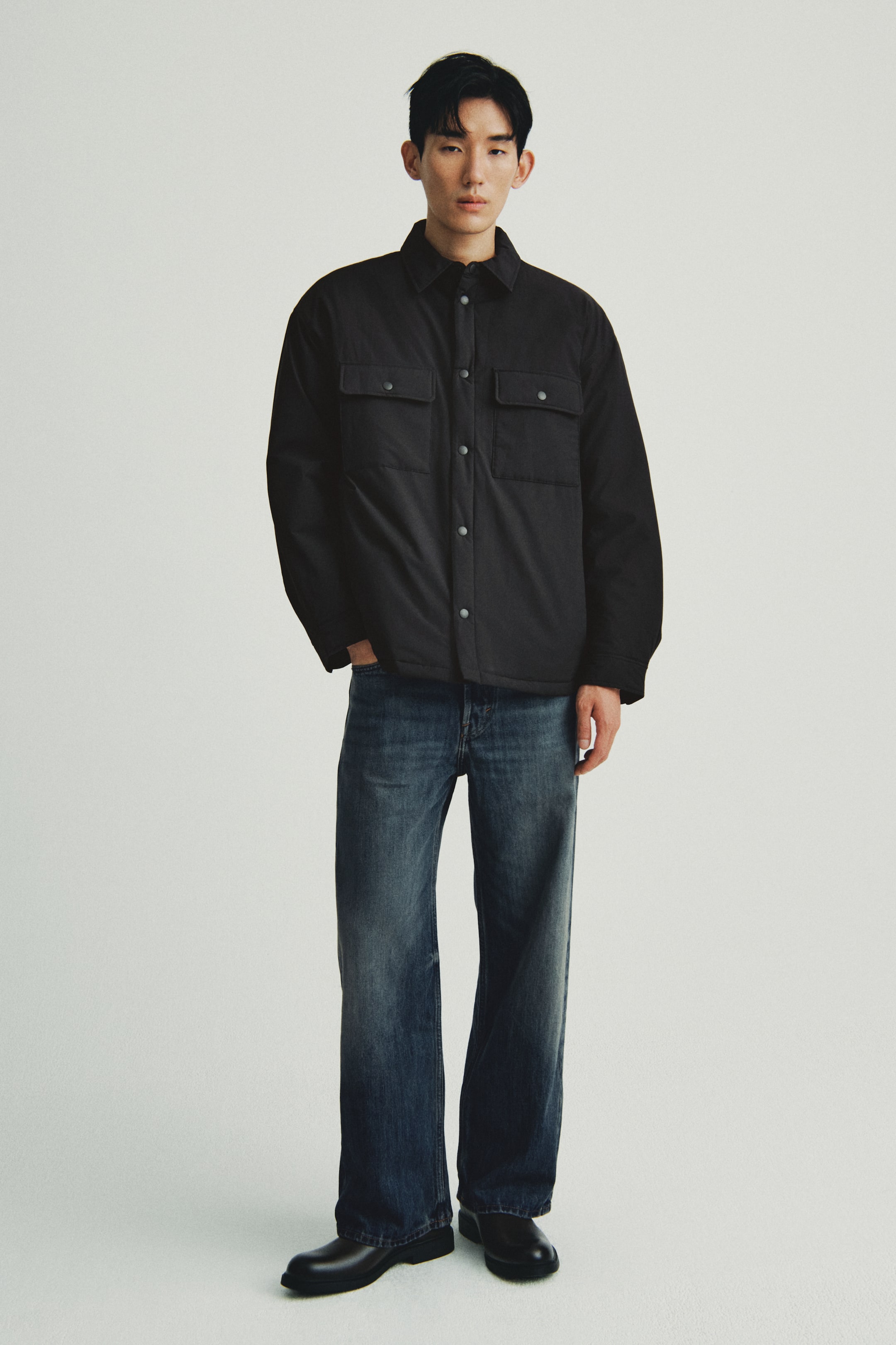 이미지 확대 보기: 검은색 샤켓이 정면으로 제시되어 있으며, 여유로운 핏과 뾰족한 칼라, 전체 스냅 버튼 앞잠금, 스냅 버튼이 있는 두 개의 플랩 가슴 포켓이 특징입니다. 또한, 다크 색상의 이너셔츠 위에 목 부분을 열어 착용하고 있습니다.