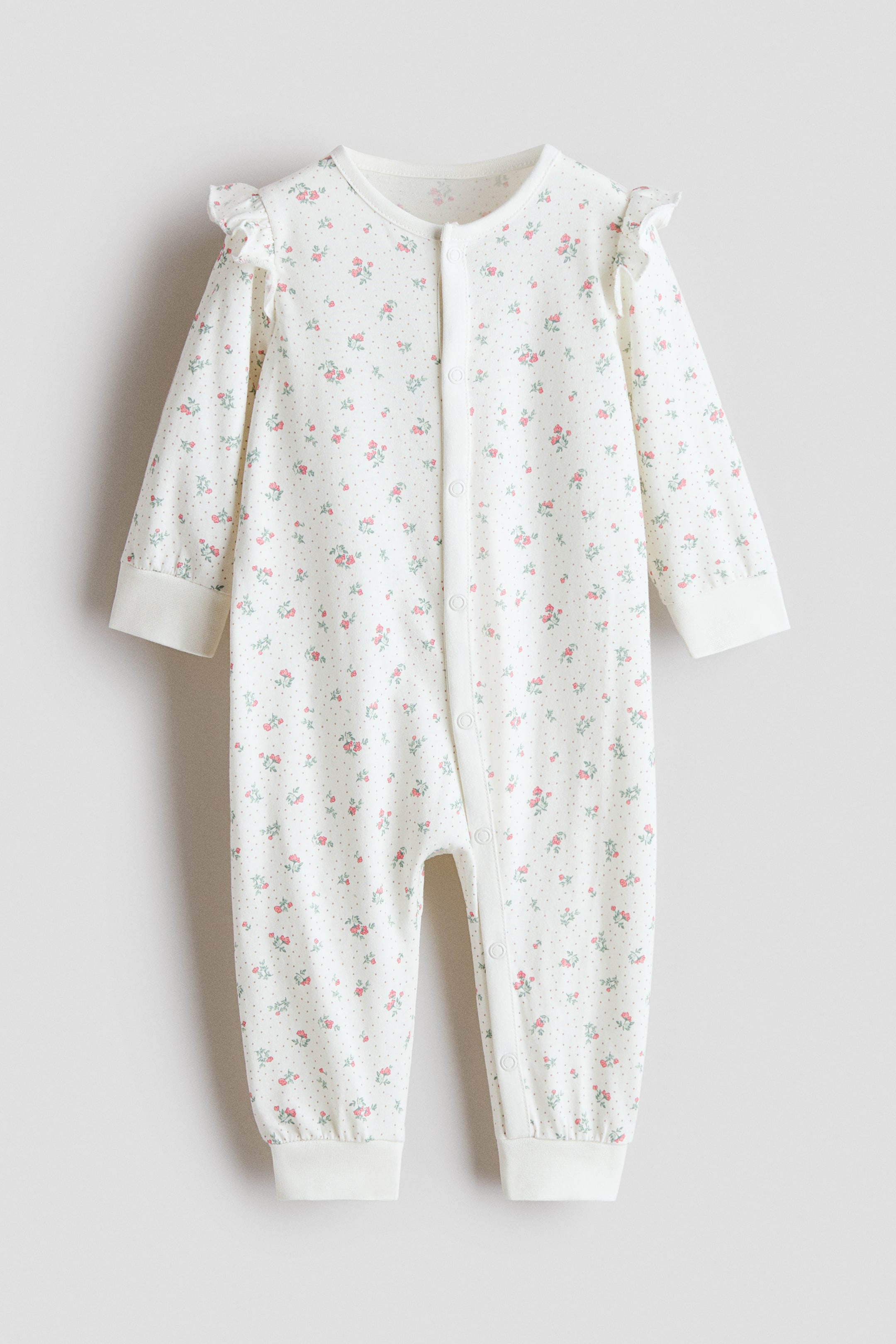 View larger image: The baby romper is a white garment adorned with a pattern of small pink roses, green leaves, and grey dots. It features ruffled trim on each shoulder, long sleeves ending in ribbed cuffs, and snap buttons running from the round neckline down the front and along one leg, terminating at ribbed ankle cuffs.