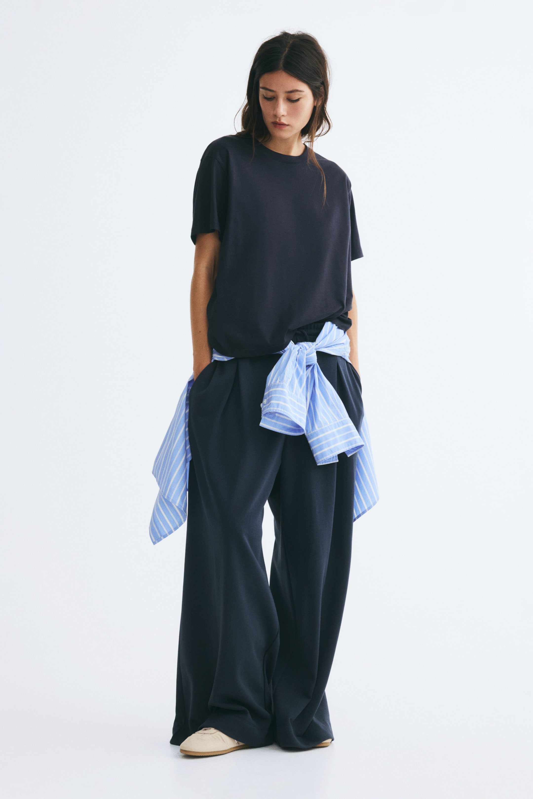 View larger image: The model is wearing a dark navy, straight-cut T-shirt with a ribbed crew neckline and subtly dropped shoulders. The T-shirt is styled with matching wide-leg trousers and a blue striped shirt tied around the waist.