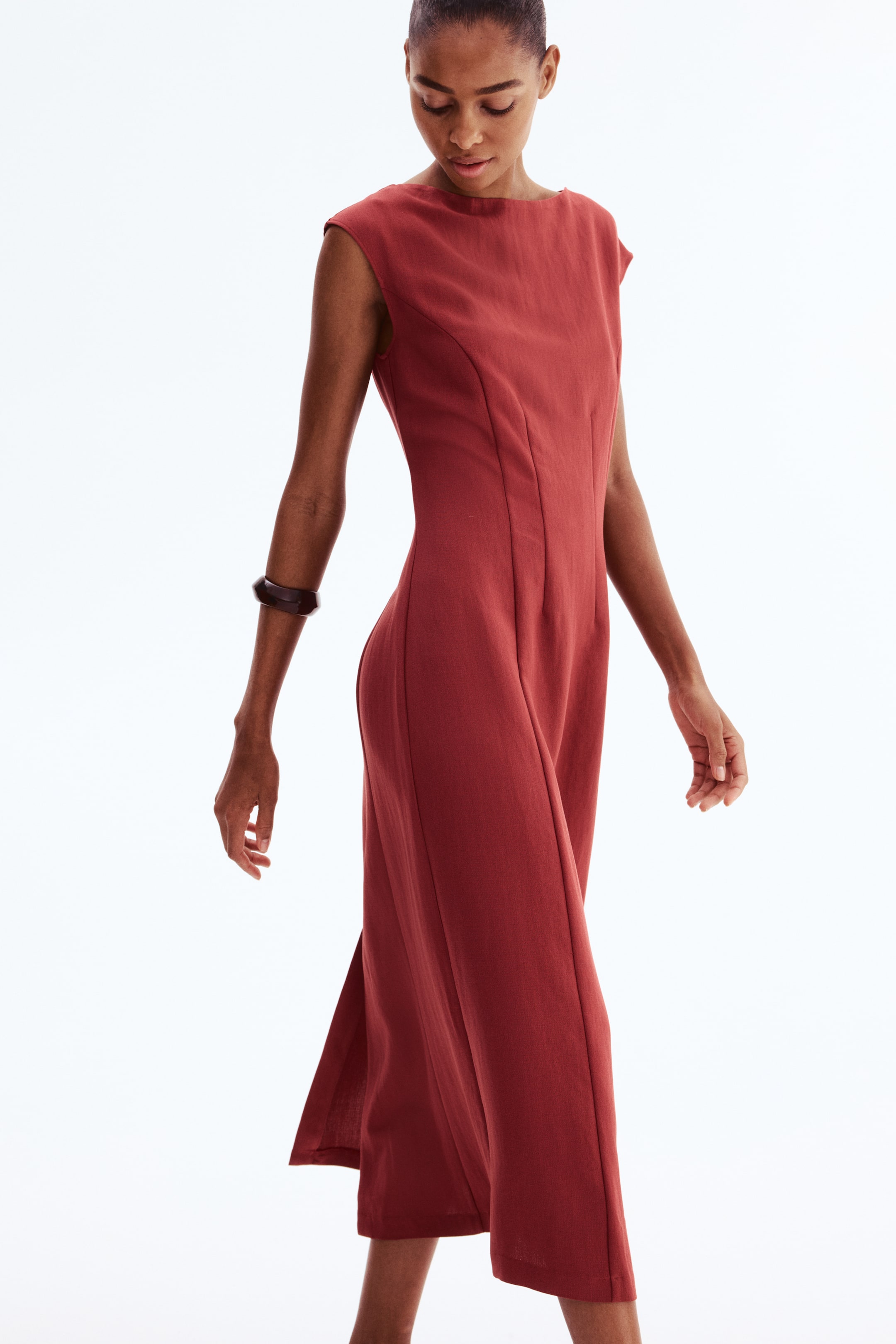 View larger image: A rust-coloured midi dress featuring a boat neckline, cap sleeves and vertical seams that shape the bodice and waist. It falls to a midi length with side slits near the hem and is presented from a dynamic side angle, highlighting the fitted bodice and flowing skirt.