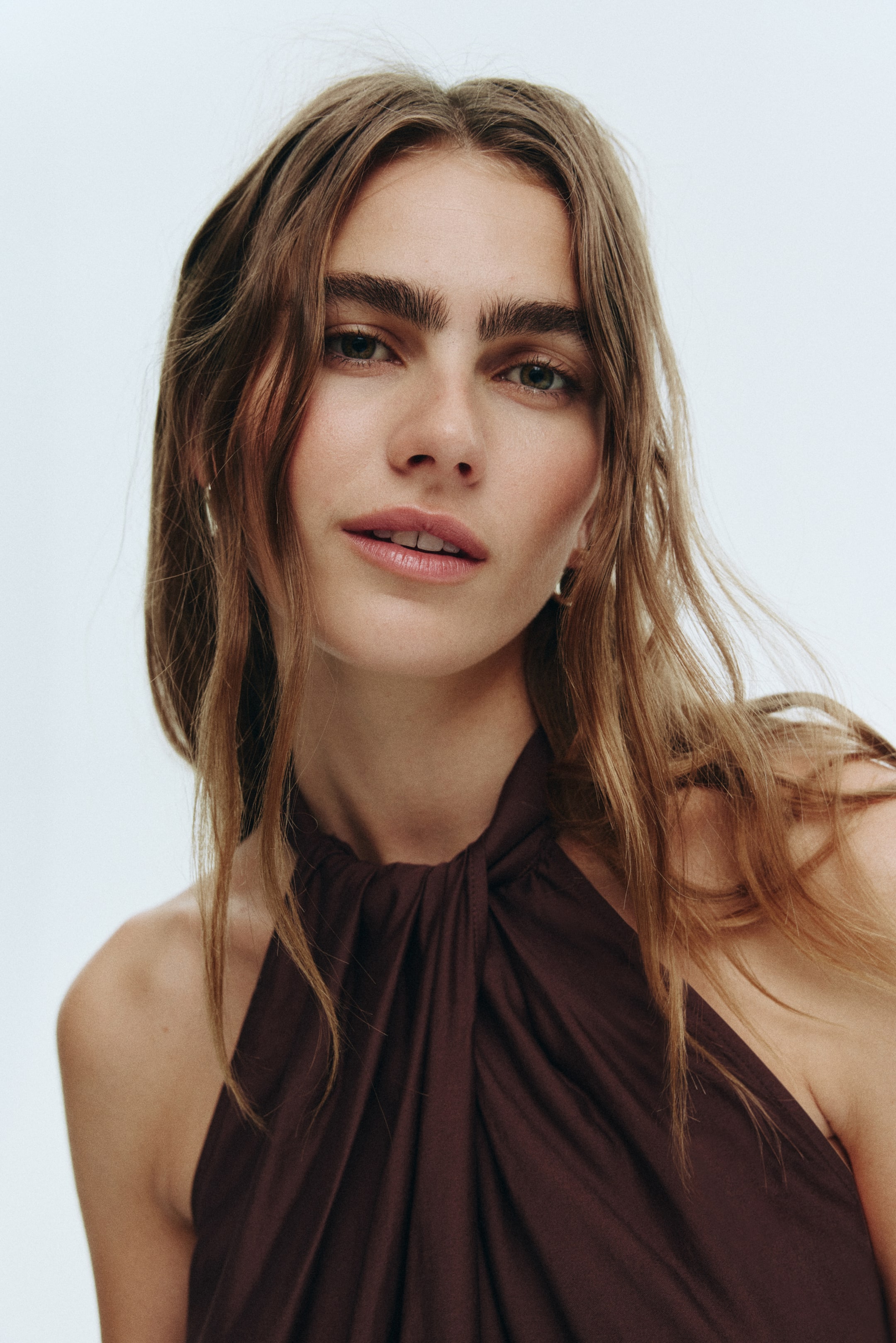 View larger image: A dark brown halter top is featuring a twisted knot detail at the center of the neckline. The top is seen from a front-facing perspective, slightly angled to the viewer's right, with fabric softly draping from the twisted element across the chest and over the left shoulder.