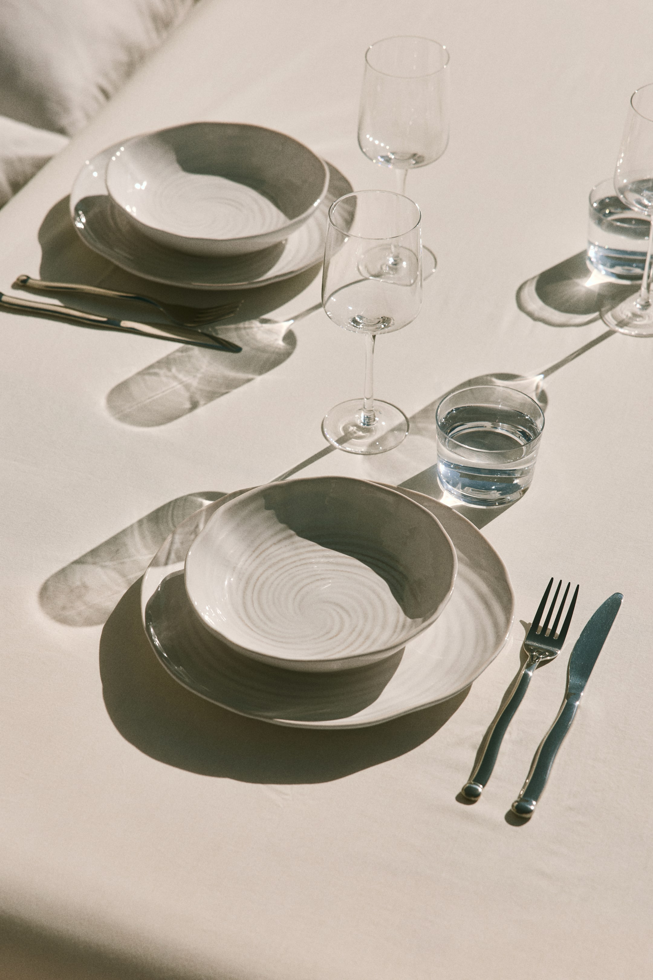 View larger image: A light-coloured ceramic dinnerware set is being presented, featuring matte off-white dinner plates with gently curved, organic edges and bowls stacked on top, which are displaying subtle spiral textures on their interior surfaces. Silverware and clear glassware are positioned alongside the settings.
