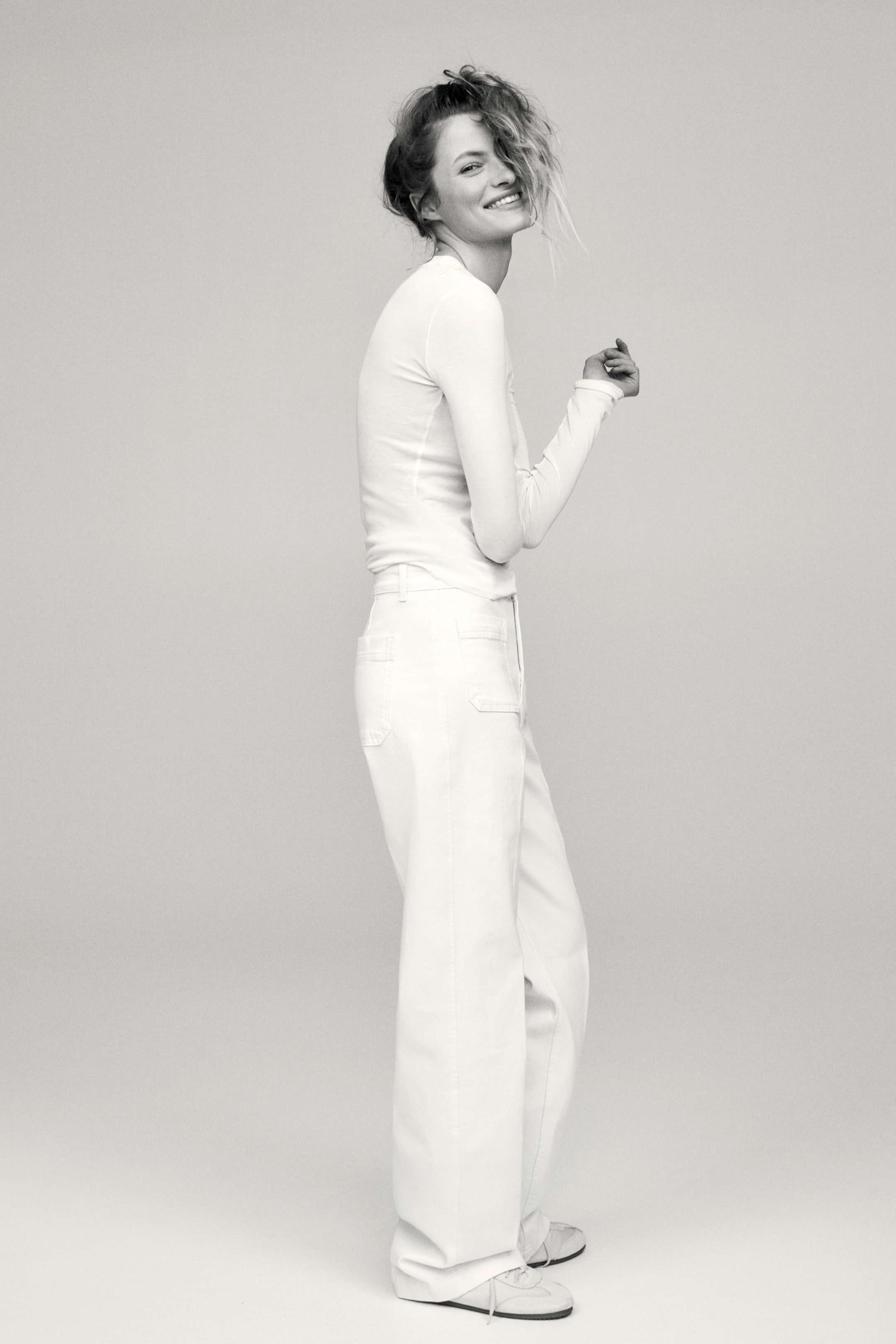 Prikaži veću sliku: Bela majica dugih rukava sa okruglim izrezom nosi se uvučena u bele pantalone sa visokim strukom i širokim nogavicama, prikazane iz bočnog ugla. Pantalone imaju vidljive bočne šavove i džepove u stilu radne odeće, ističući njihov opušten, pun dužinski kroj.