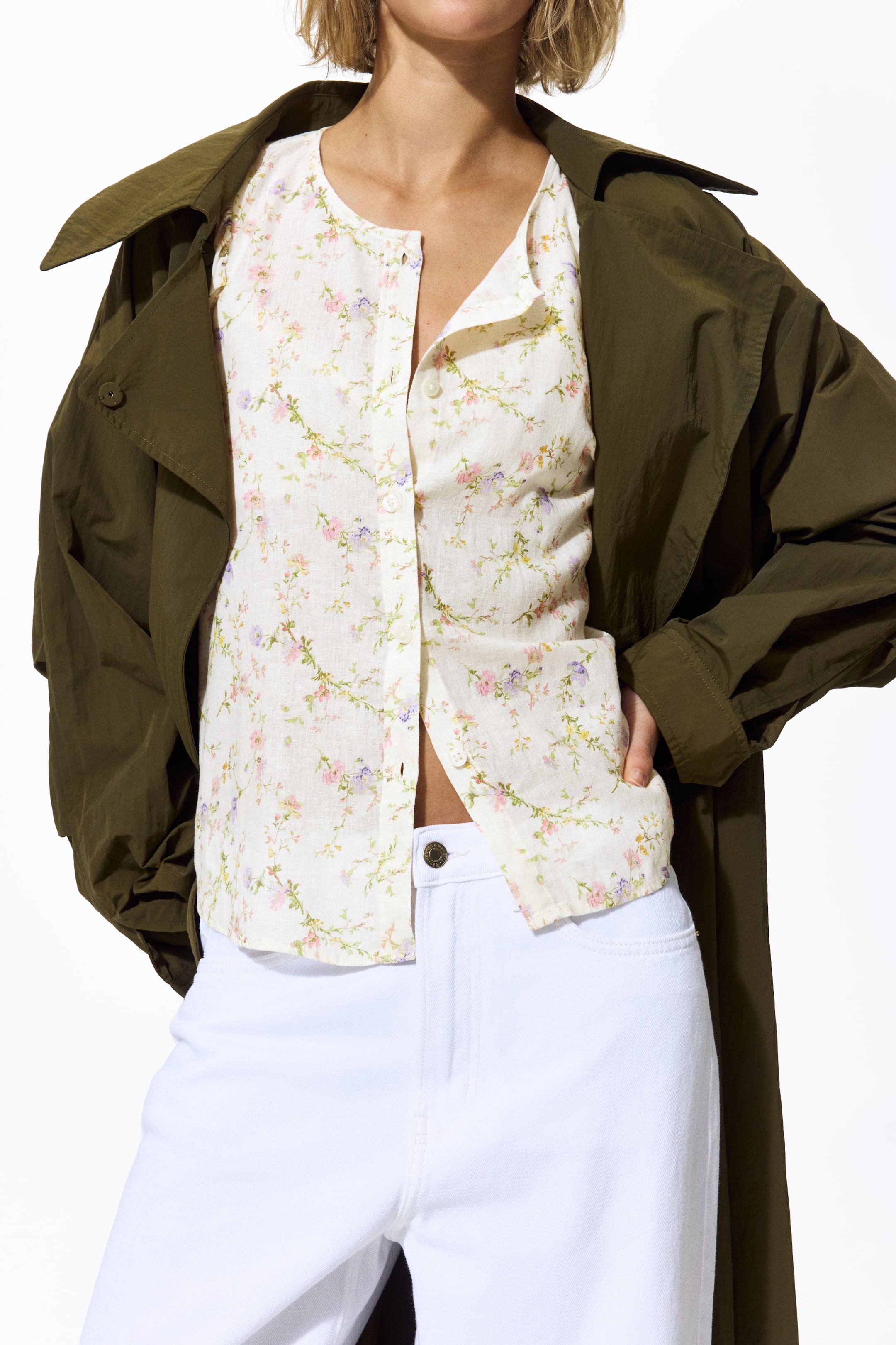 Büyük resmi görüntüle: Beyaz keten karışımlı düğmeli gömlek, yuvarlak bir yaka ve pembe, mor ve yeşil çiçeklerle sarmaşık desenine sahiptir. Ön kısmında görünen beyaz düğmeler bulunmakta olup, koyu yeşil bir trençkotun altında katmanlanmış, beyaz pantolonların üzerinde kısa görünmektedir.