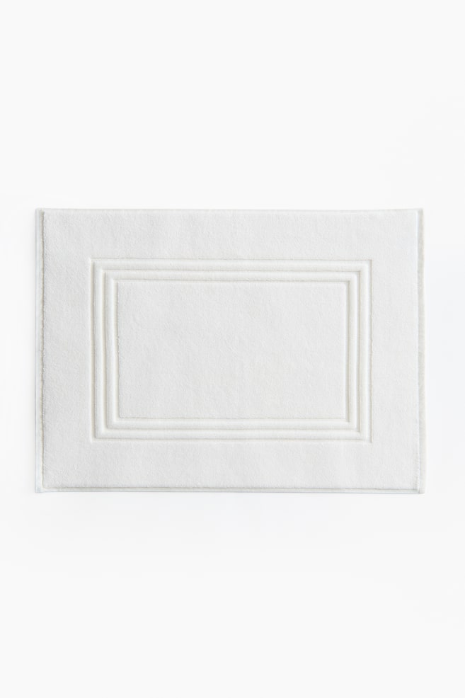 Un tapis de bain blanc rectangulaire présente une texture moelleuse et un design de bordure rectangulaire surélevée à double ligne en son centre. - 1