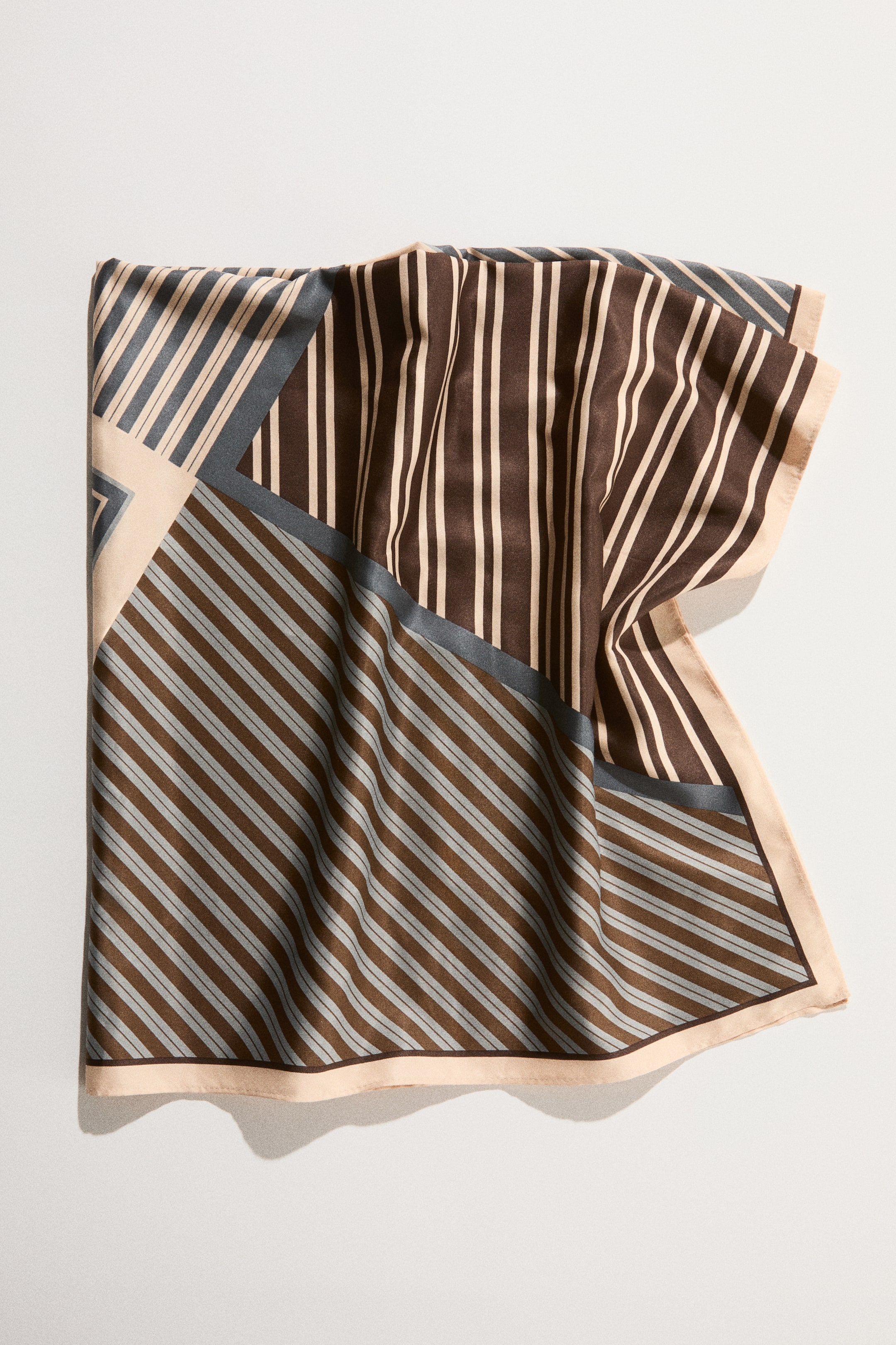 View larger image: A scarf in a soft weave features a printed geometric pattern, presented with gentle folds. It displays sections of vertical and diagonal stripes in brown, light beige, and grey, separated by solid blocks, and is edged with a light beige border.
