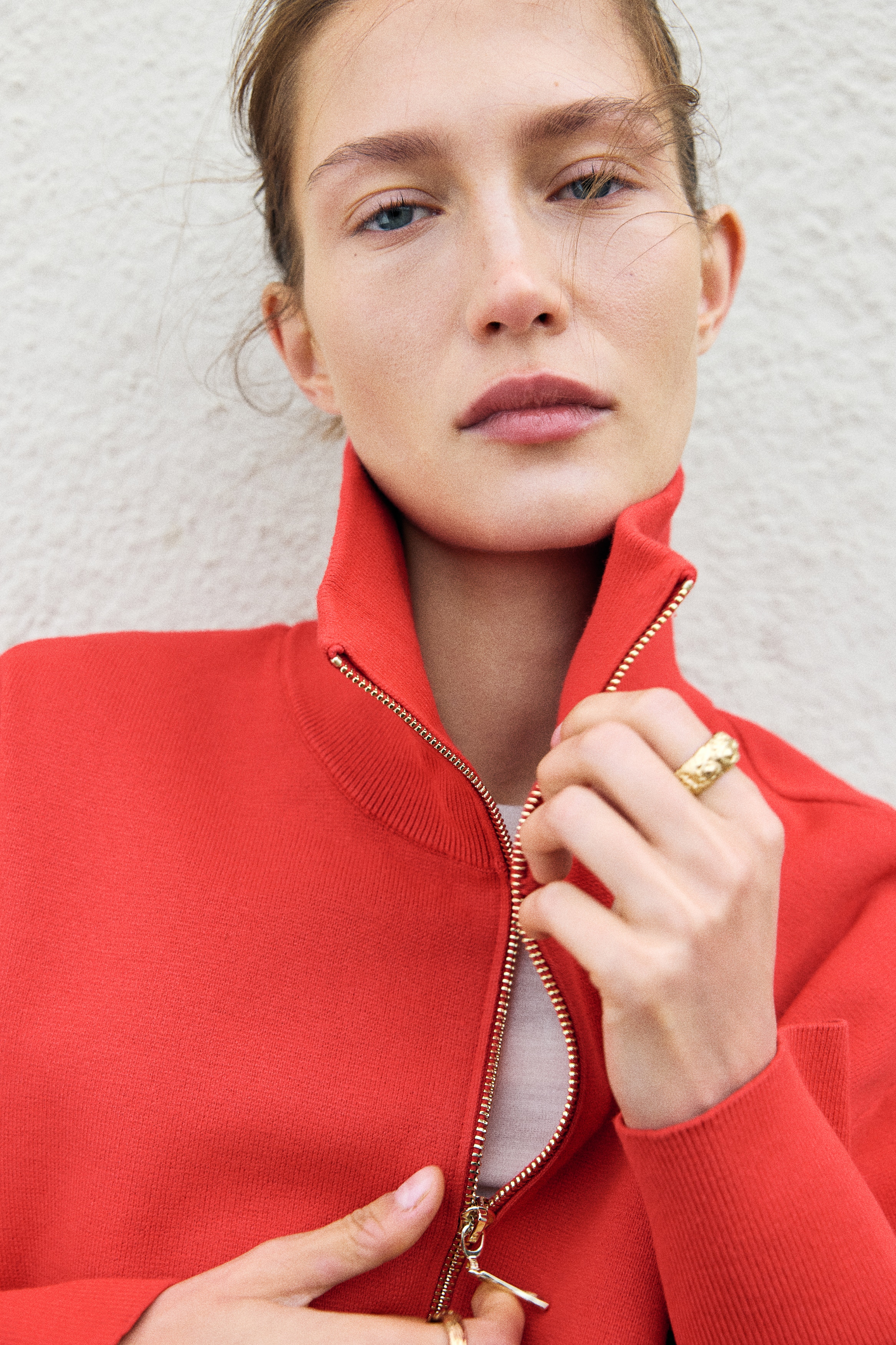 A red fine-knit sweater is shown from the front, featuring a high neck with a gold-tone half zip. The sweater has dropped shoulders, long sleeves with wide ribbed cuffs, and a matching ribbed hem.
