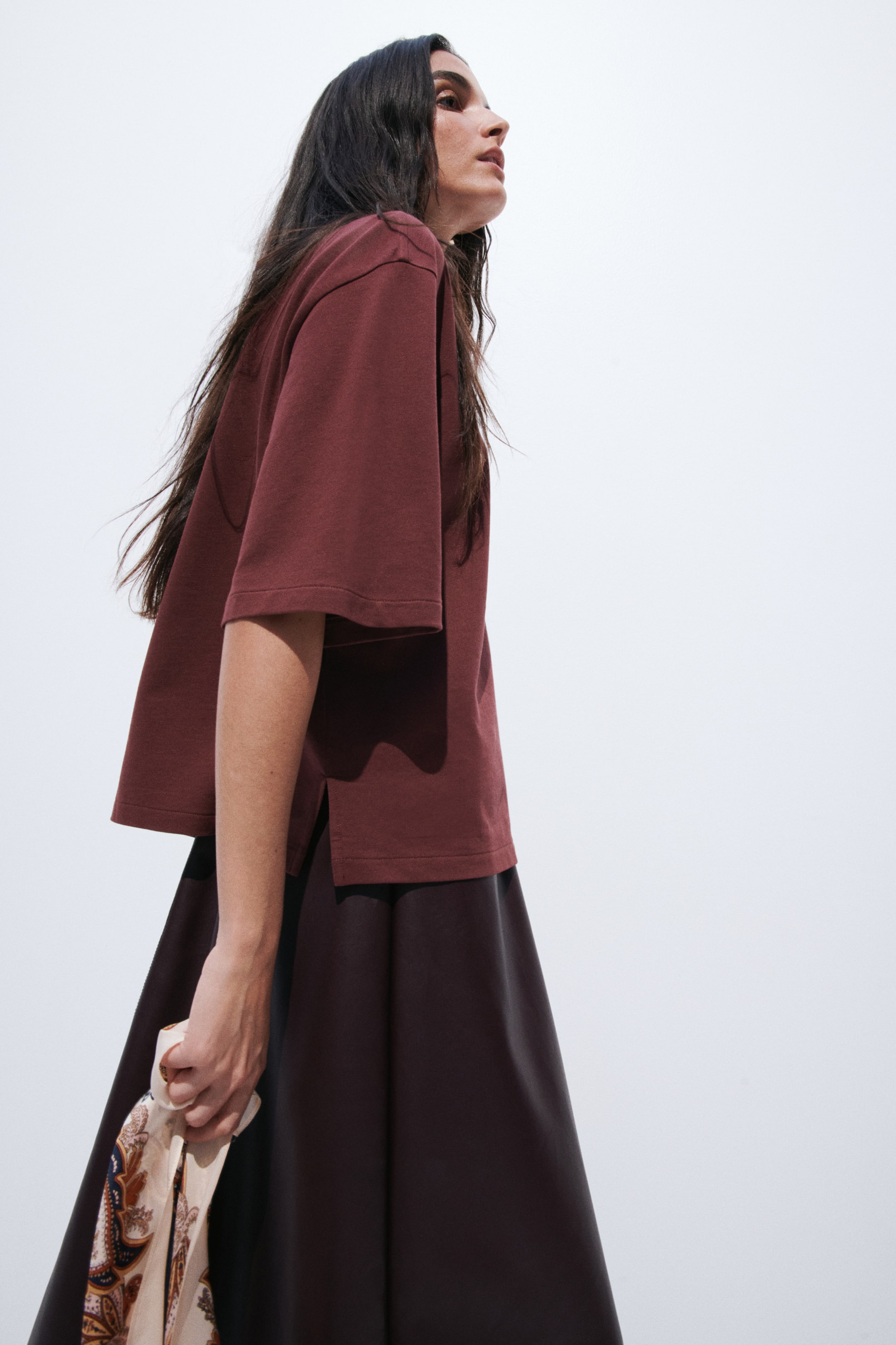View larger image: A dark reddish-brown short-sleeved top is being worn, facing towards the right, showcasing its loose, boxy fit. It features wide sleeves ending above the elbow and a straight hem with side slits, layering over a dark skirt.