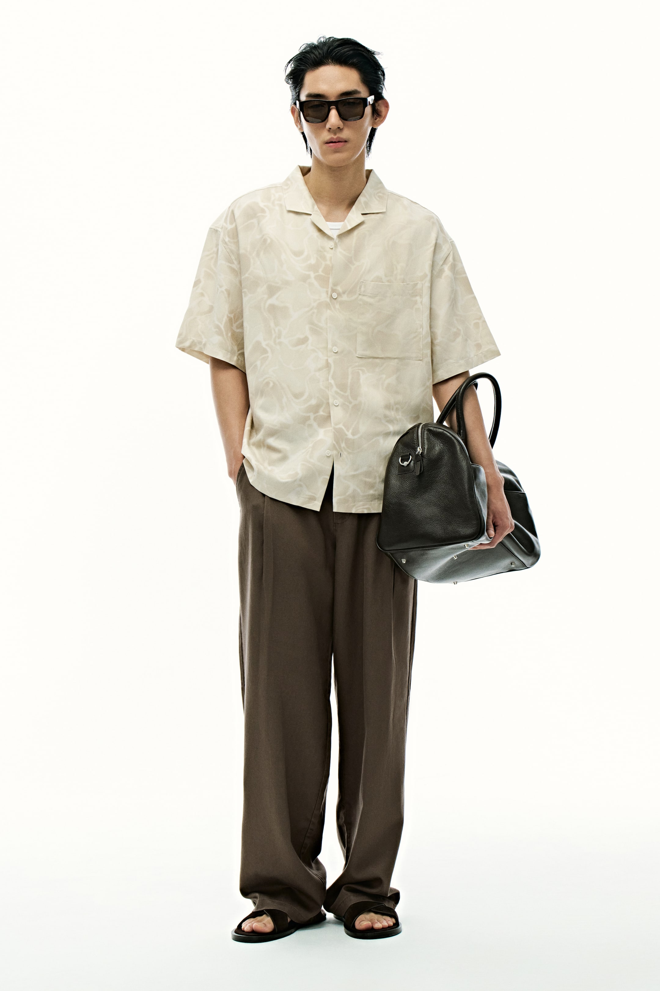 View larger image: A light beige short-sleeved camp collar shirt, featuring a subtle abstract pattern and a chest pocket, is being presented facing forward, styled with dark brown wide-leg pleated trousers and dark open-toed sandals. A dark leather duffel bag is being held in one hand, while dark rectangular sunglasses are being worn.