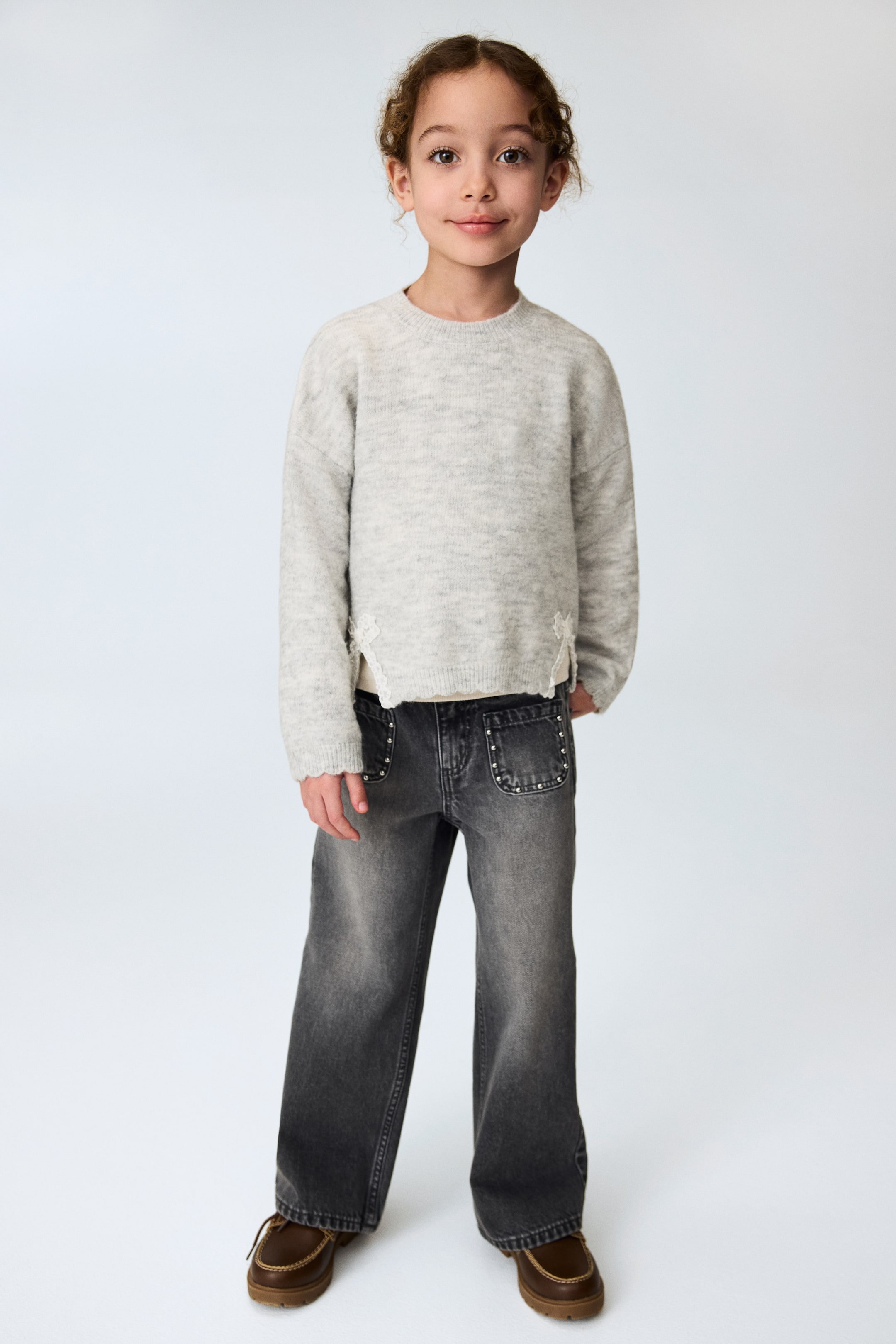 View larger image: A light grey heathered knit jumper is being displayed from a front view, featuring a crew neck, scalloped cuffs, and a scalloped hem with white lace detailing at the side slits. It is styled over dark grey wide-leg jeans that are showcasing studded front pockets and a faded finish, with brown moccasin-style shoes completing the look.