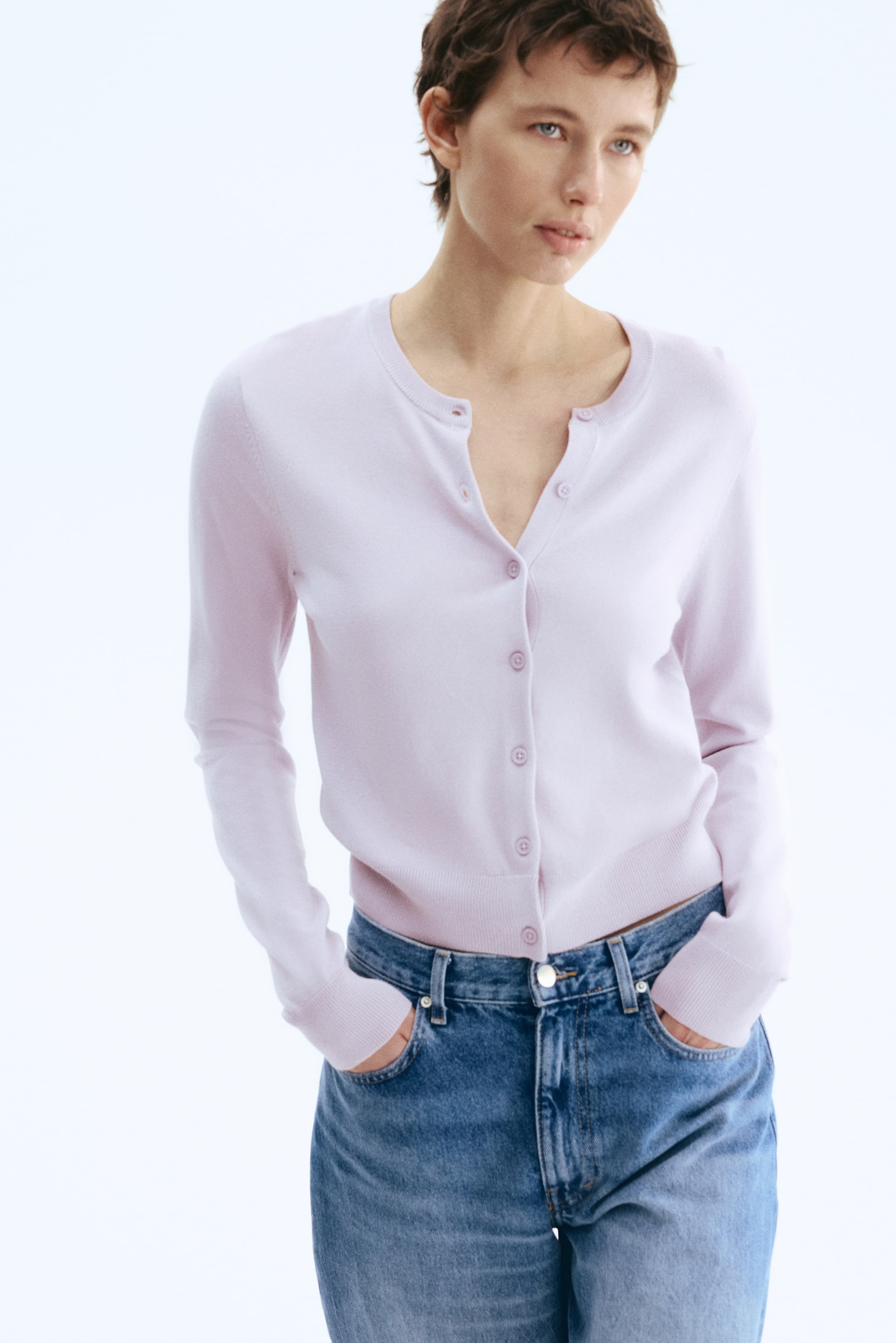 Vezi imaginea mai mare: Un cardigan tricotat fin, de culoare roz deschis, cu decolteu rotund, nasturi pe partea din față și manșete și tivuri cu nervuri, este purtat de model. Modelul poartă cardiganul parțial descheiat peste blugi albaștri, cu mâinile în buzunarele blugilor.