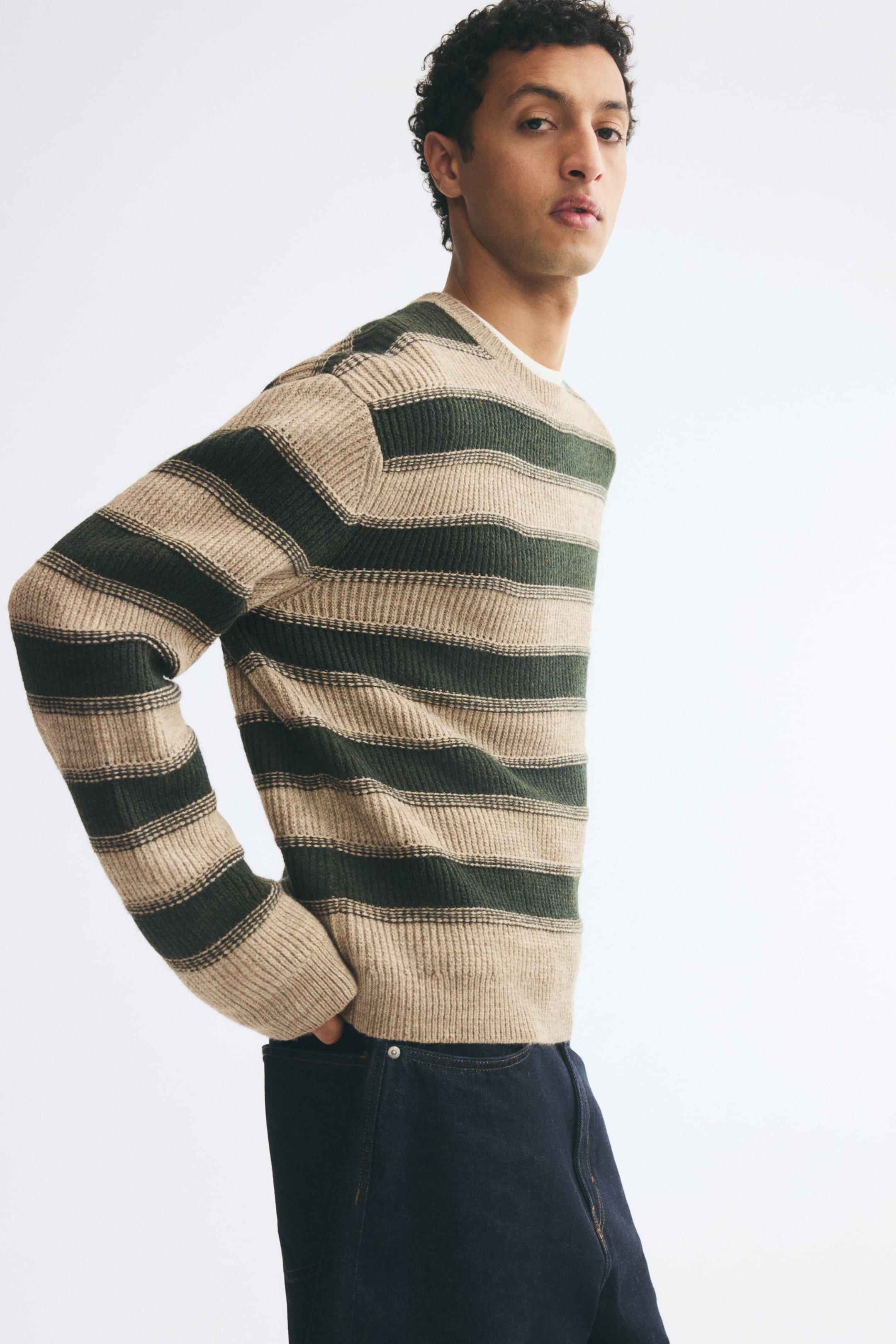 Visualizza immagine più grande: Un maglione a coste a righe in beige e verde scuro indossato dalla modella, caratterizzato da girocollo, spalle scese e finiture a coste su polsini e orlo. Il maglione è abbinato a jeans blu scuro, con la modella che posa una mano vicino alla tasca posteriore.