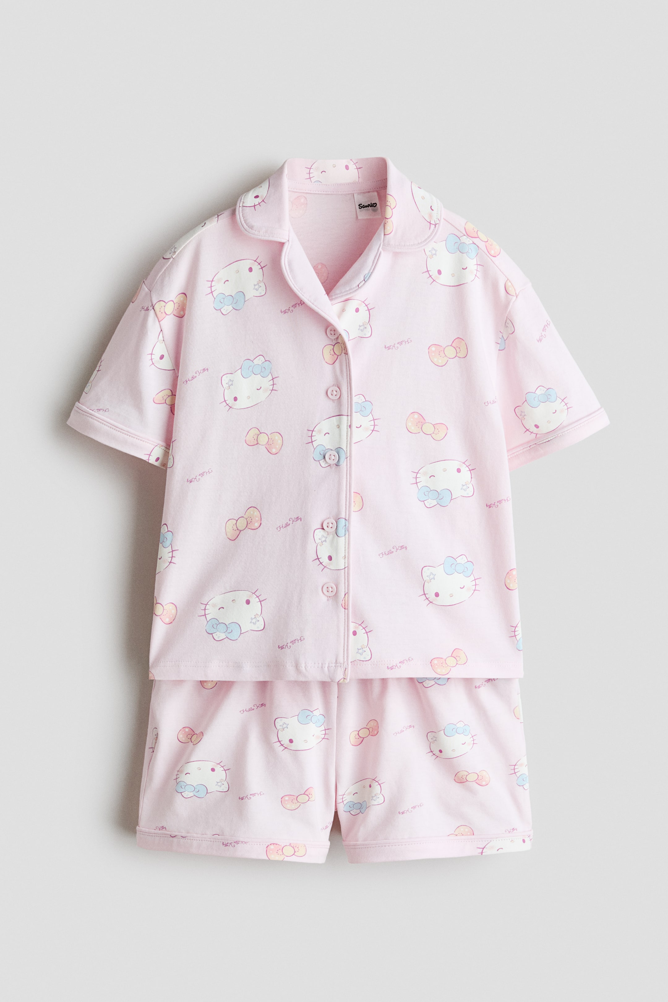 View larger image: A pink two-piece pyjama set is facing front. The short-sleeved top features notch lapels and a front button-up closure, showcasing a pattern of Hello Kitty faces with bows and individual bows. The matching shorts display the same pattern.