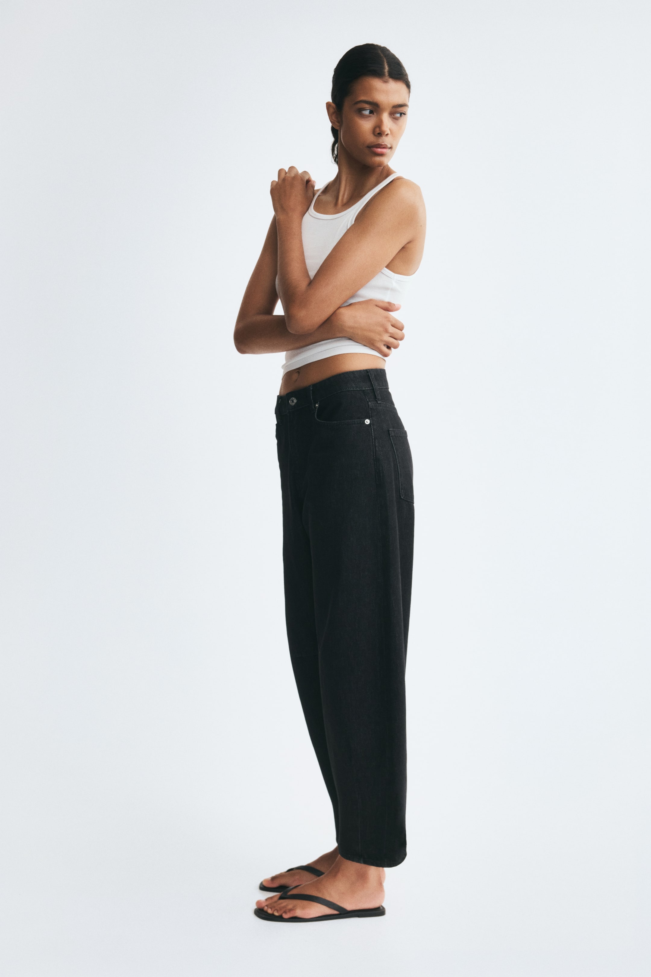Xem ảnh lớn hơn: Người mẫu đang mặc quần jeans denim đen cạp cao với phom ống cong, rộng và thuôn gọn ở mắt cá chân. Quần jeans được phối cùng áo ba lỗ gân màu trắng và dép sandal bệt màu đen.