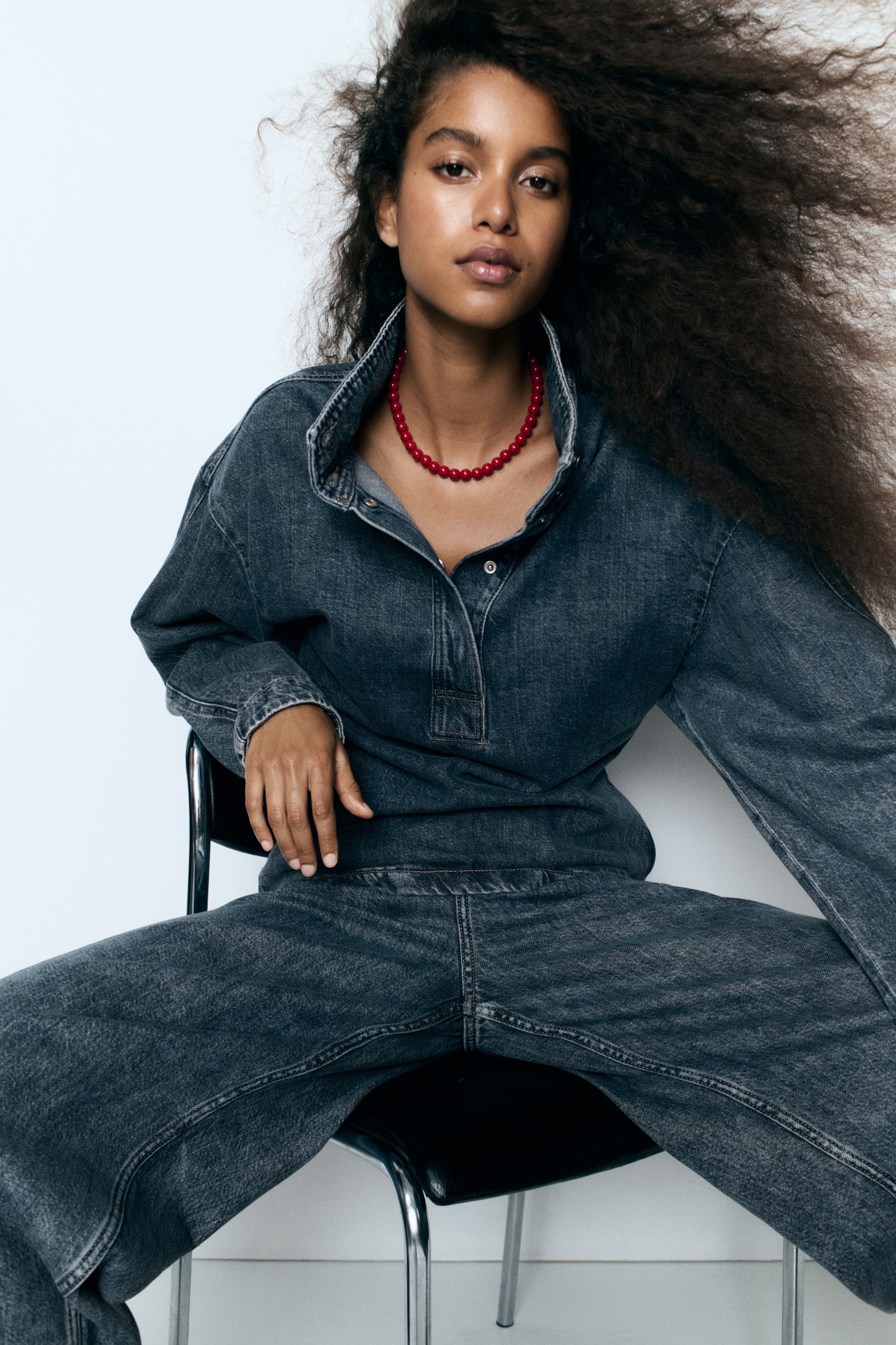Vezi imaginea mai mare: O cămașă popover din denim gri închis, cu guler tunică cu nasturi, fenta parțial deschisă și umeri căzuți, este purtată de model. Cămașa este asortată cu blugi din denim închis la culoare și un colier roșu cu mărgele.
