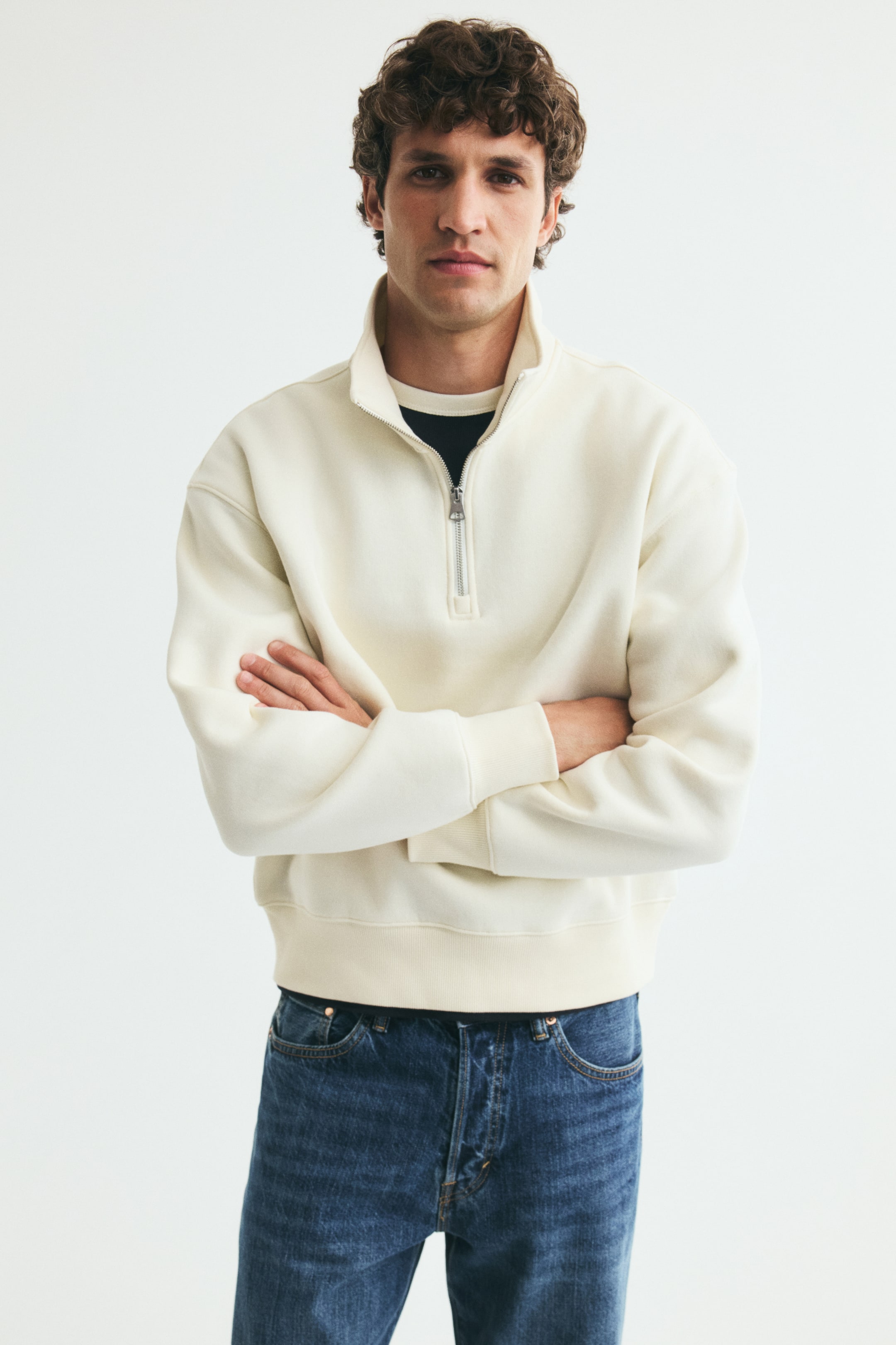 View larger image: An off-white half-zip sweatshirt is being worn by the model, who is standing with arms crossed. The sweater features a stand collar, dropped shoulders, and ribbed cuffs and hem, and is styled over a dark crew-neck T-shirt and blue jeans.