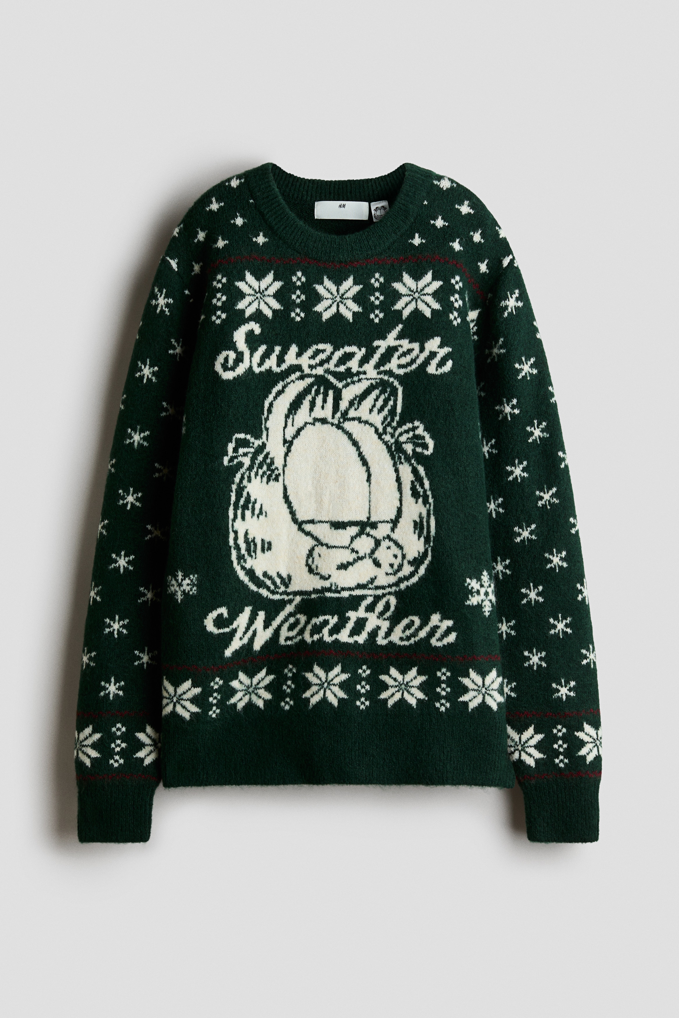 A dark green jacquard-knit sweater is shown from the front, featuring a round rib-trimmed neckline, long sleeves, and a ribbed hem. The front displays a large white graphic of the cartoon character Garfield with the cursive words "Sweater Weather," bordered by horizontal bands of white snowflake patterns and thin red stripes.