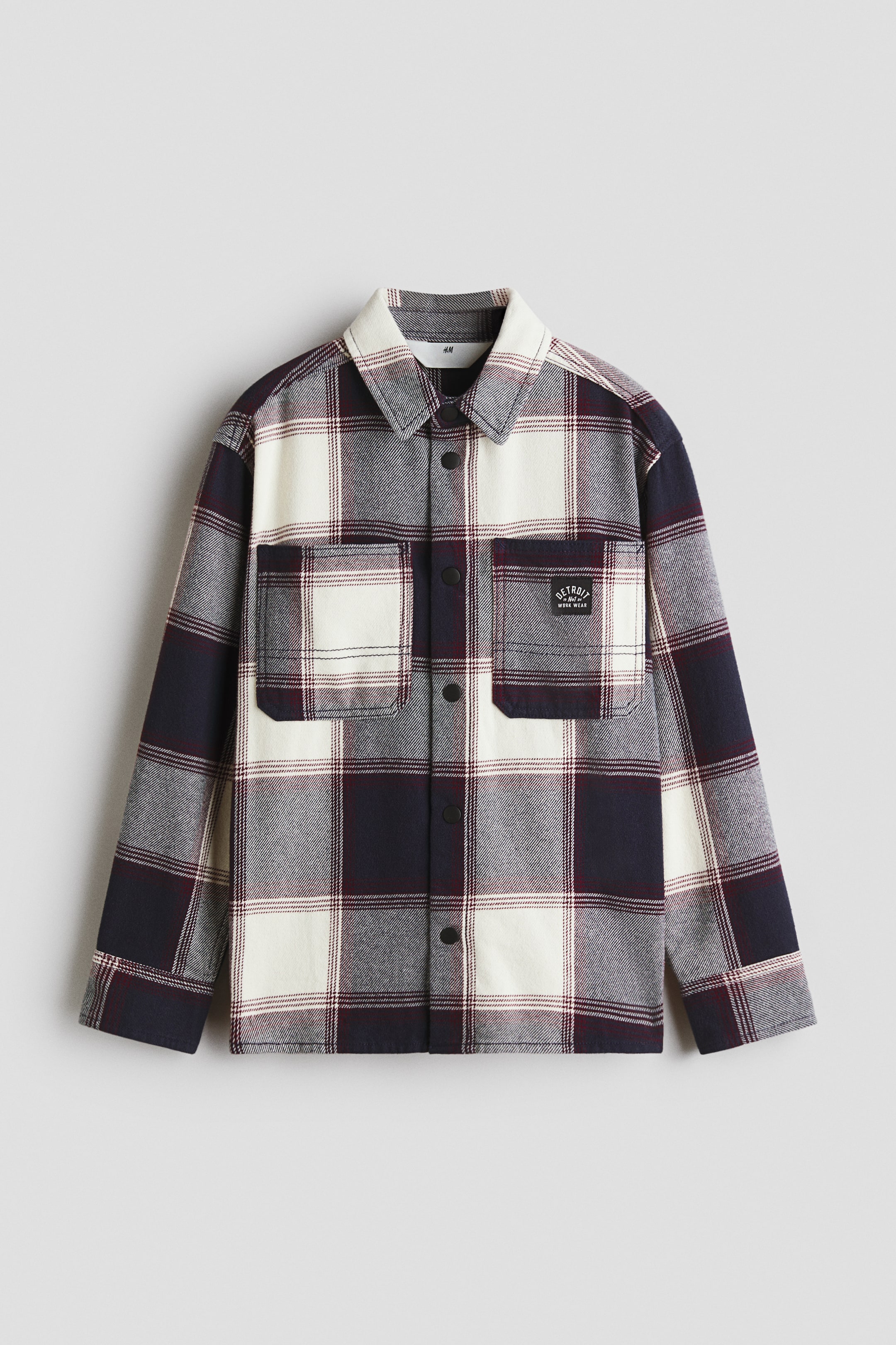 View larger image: A check overshirt is showing its front, featuring a pattern of white, medium grey, dark navy, and maroon squares. It is designed with a classic collar, a button-down front closure with dark buttons, and two chest flap pockets, one displaying a small black patch label.