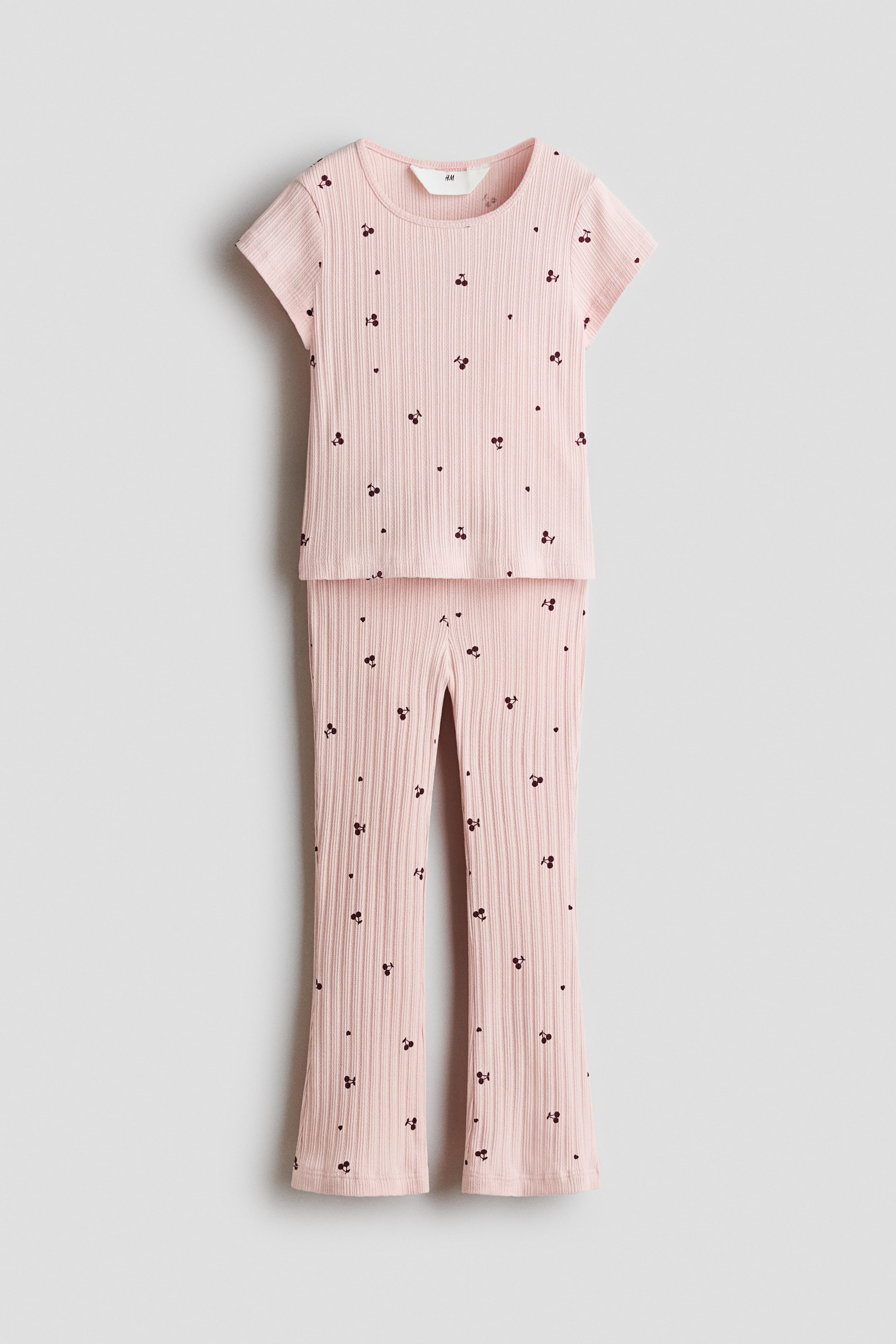 Ver imagen más grande: Conjunto de dos piezas en color rosa claro acanalado que incluye un top de manga corta y pantalones acampanados, ambos con un estampado completo de pequeñas cerezas oscuras y diminutos corazones oscuros.