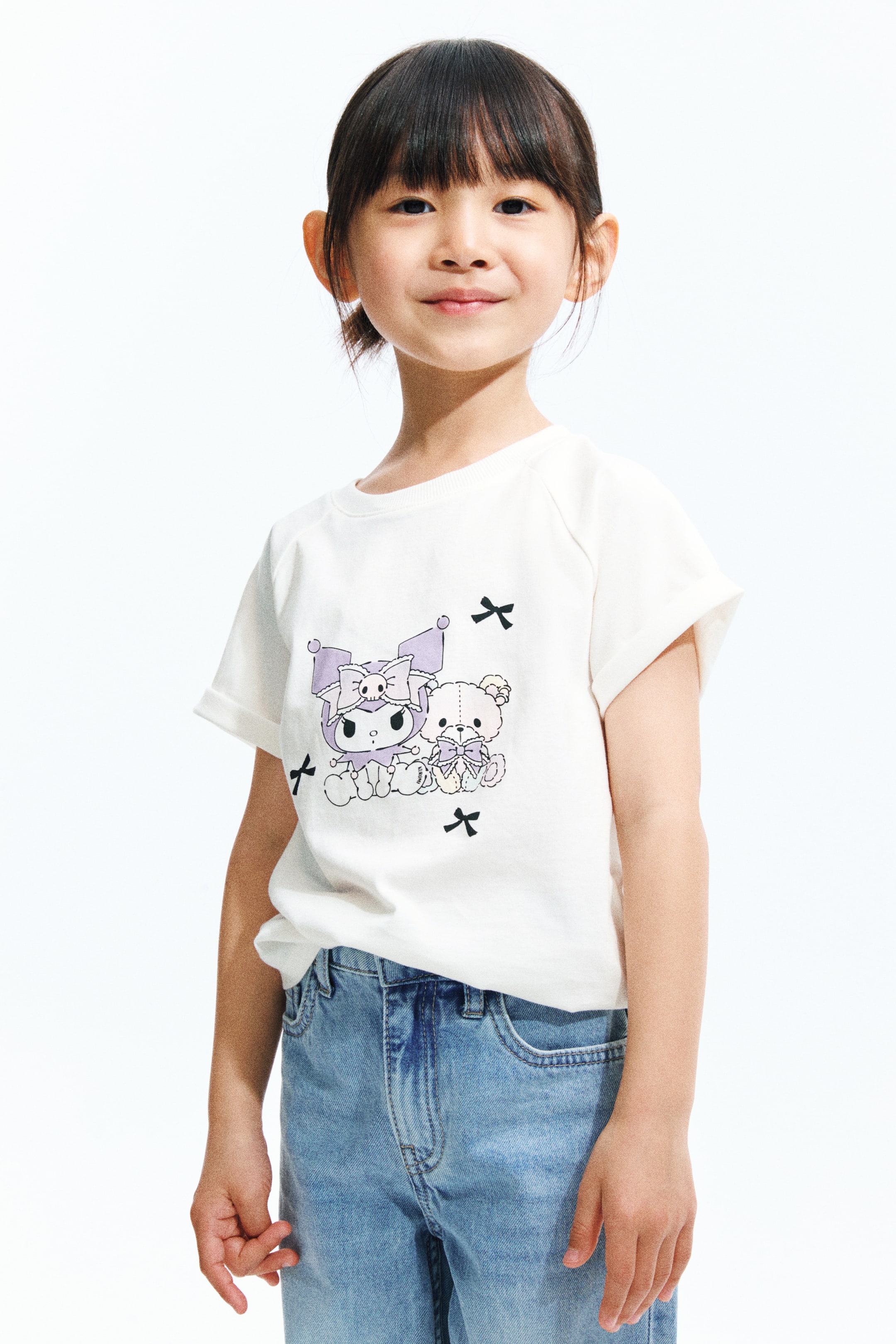 拡大画像を表示: 白いTシャツは、紫色のキャラクターとテディベアのグラフィックプリントがフロントに施され、黒いリボンでアクセントが付けられています。ショートスリーブはロールアップされており、リブ付きのクルーネックデザインです。ライトブルーのジーンズに部分的にタックインされています。