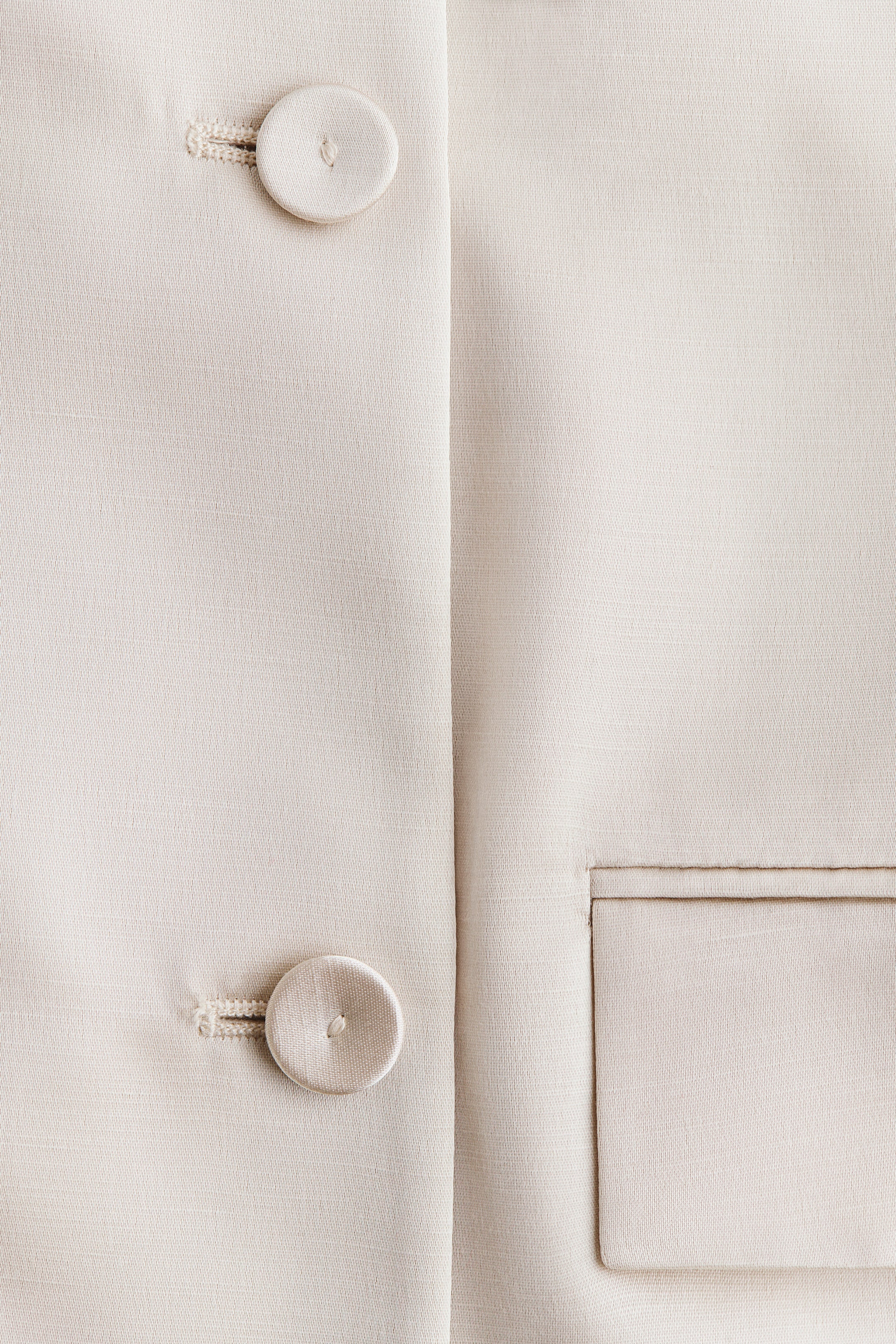 View larger image: A light-colored woven blazer dress is displaying two round, fabric-covered buttons fastened with thread loops on its front left, and a jetted pocket with a rectangular flap on its front right.