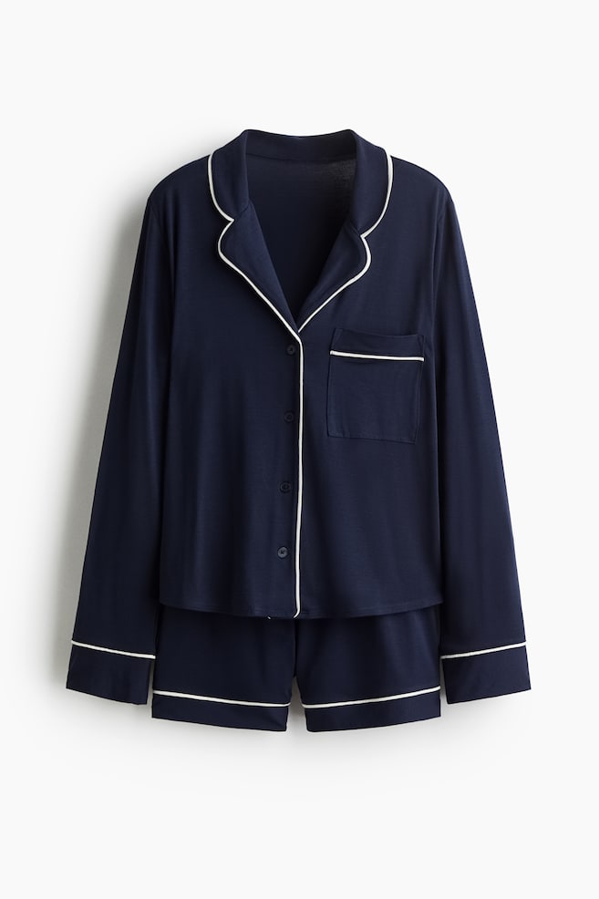 A navy pyjama set is being presented from the front, consisting of a long-sleeved shirt and shorts. The shirt is featuring a resort collar, a buttoned front placket, an open chest pocket on the left, and long sleeves with cuffs, all accented with white piping. The matching shorts are displaying white piping along their hems. - 1