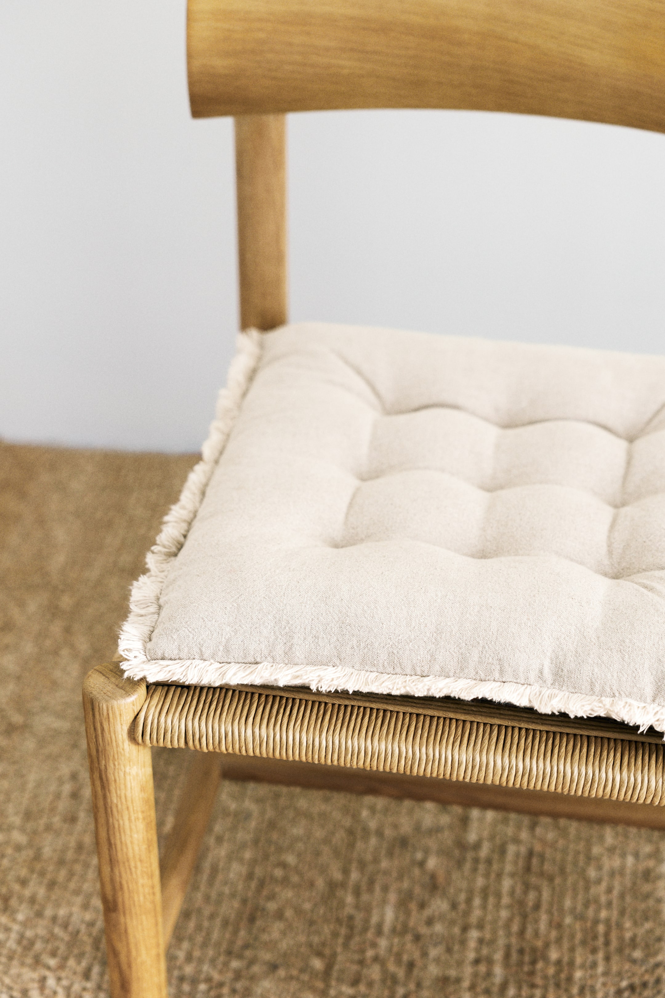 View larger image: A padded seat cushion is shown from a front-side angle, featuring a light beige linen and cotton weave, thick padding, evenly spaced quilt knots across its surface, and frayed edges around its perimeter.