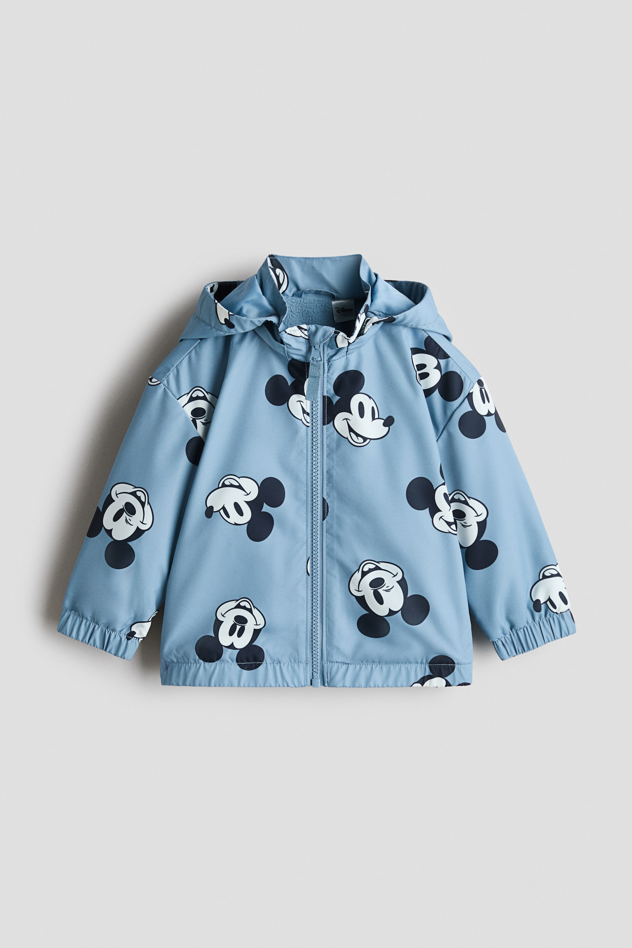 Ampliar la imagen: Una chaqueta con capucha de color azul claro para niño presenta el FRENTE, con un patrón repetitivo de cabezas de Mickey Mouse en blanco y negro. Está diseñada con un cierre frontal de largo completo, puños elásticos y un dobladillo elástico.