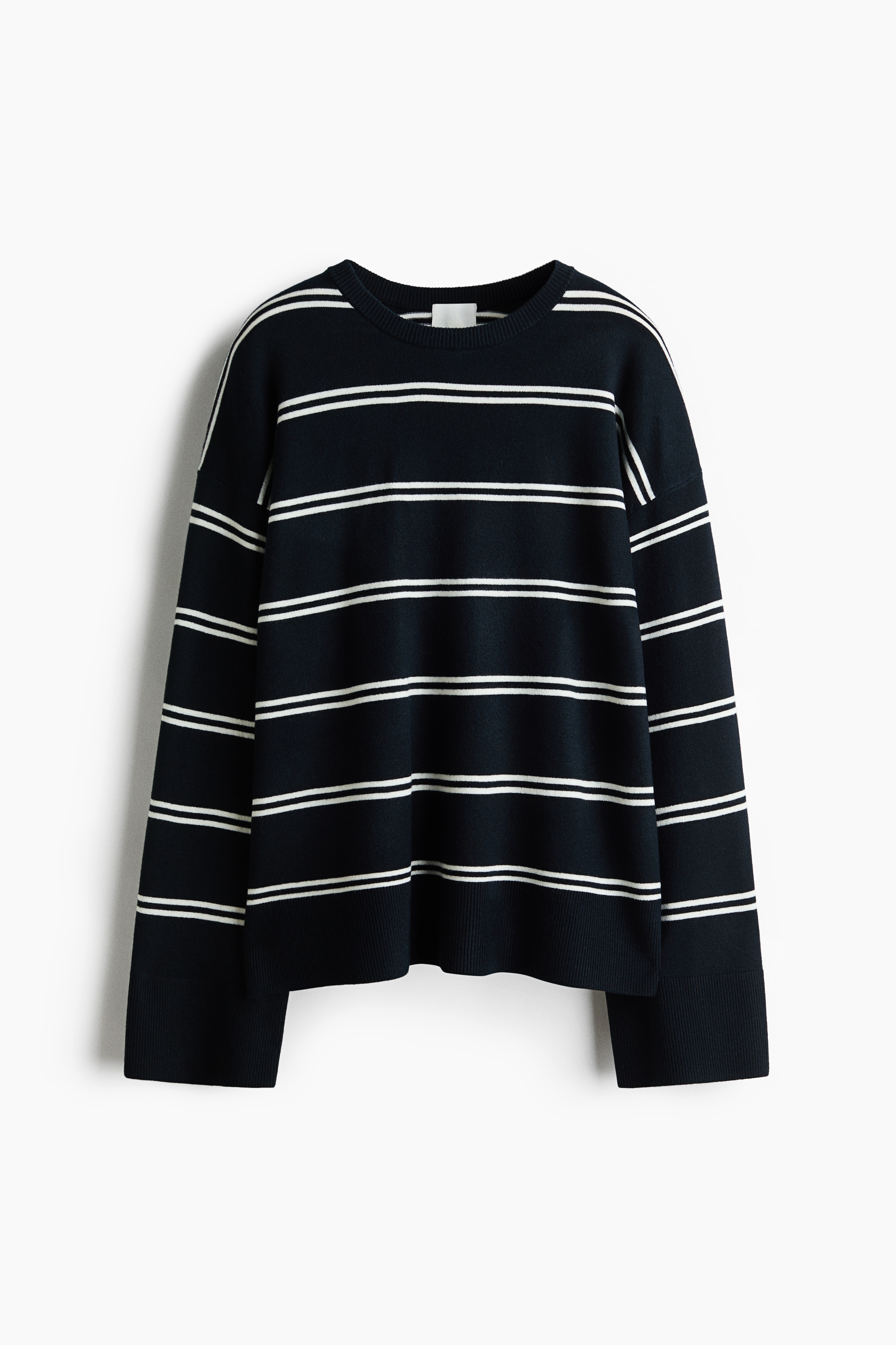An oversized knit sweater in dark navy blue with horizontal white double-stripes is being shown from a side angle facing left. The sweater is featuring a round ribbed neckline, dropped shoulders, and long sleeves with wide ribbed cuffs.