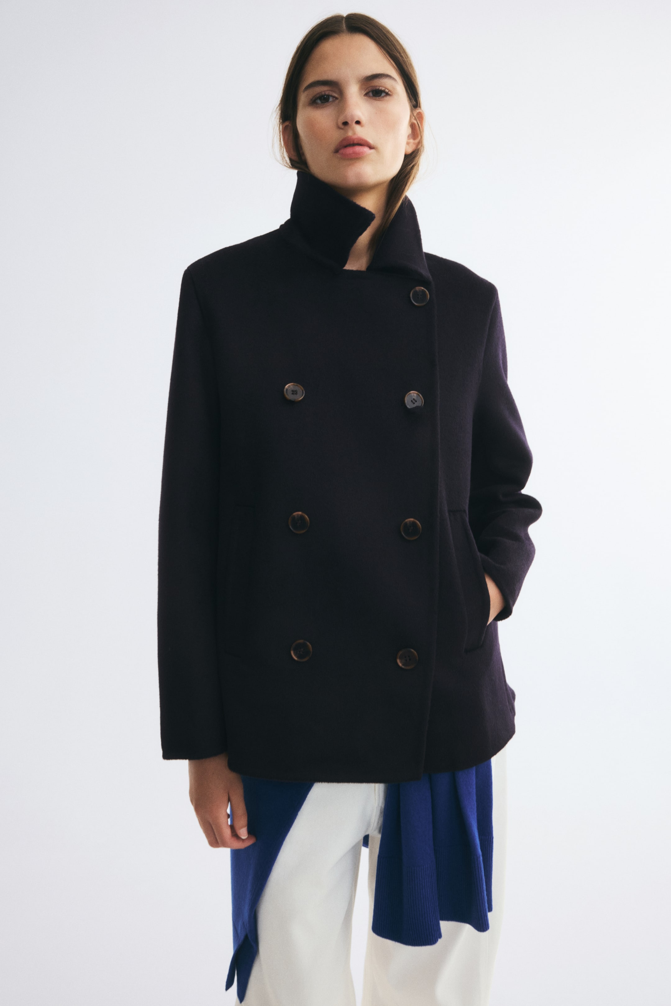 Visualizza immagine più grande: Un cappotto corto doppiopetto in una tonalità blu navy scuro, realizzato in un misto lana infeltrita, è indossato dalla modella, che tiene una mano in una tasca a filetto laterale. Il cappotto presenta un colletto alto risvoltato e chiusura frontale con bottoni, ed è abbinato a pantaloni bianchi e a un maglione blu annodato in vita.