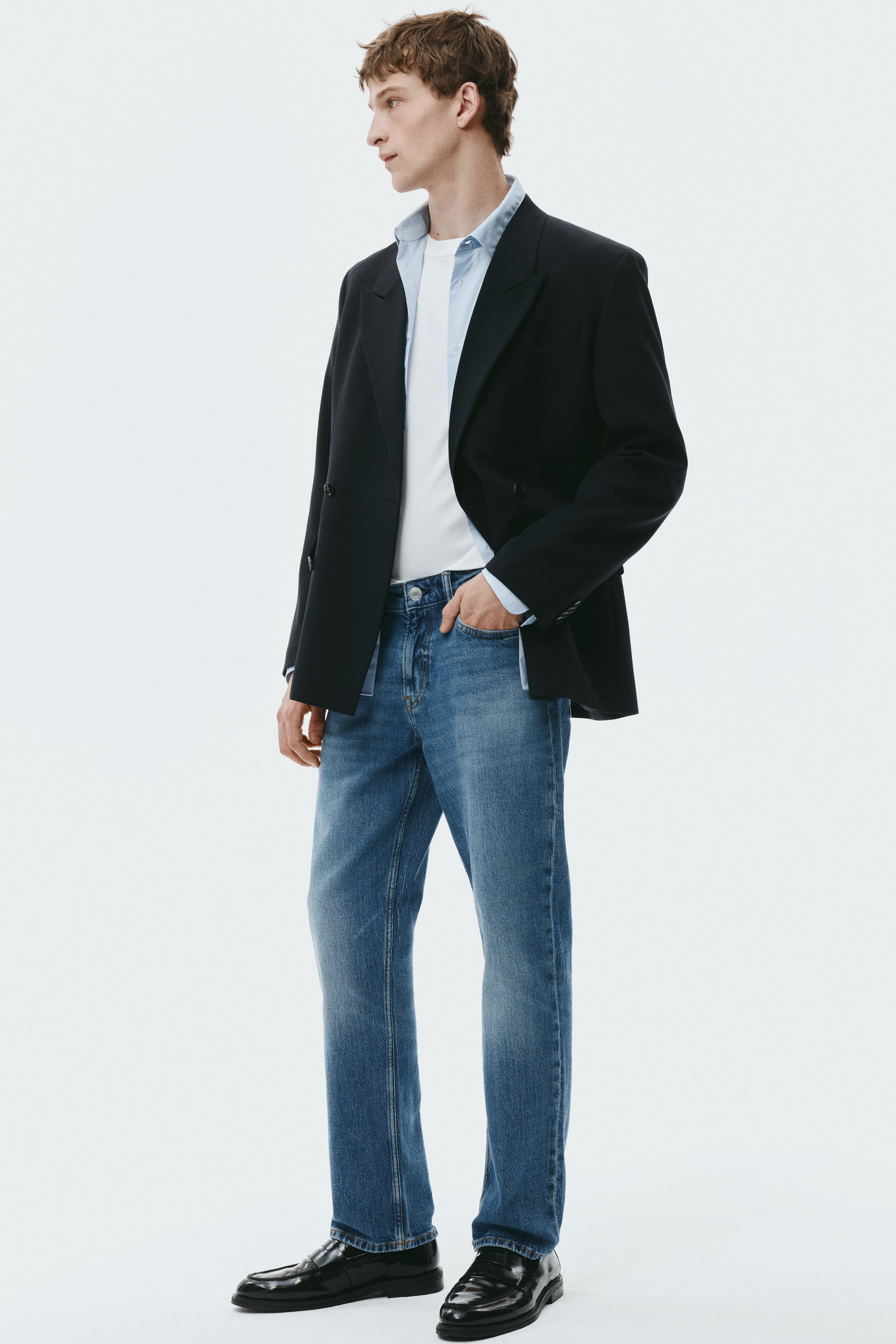 View larger image: A black double-breasted blazer with notch lapels is being worn open over a light blue collared shirt and a white crew-neck t-shirt. Medium-wash blue straight-leg jeans are being styled with black polished loafers, all presented from a three-quarter angle.