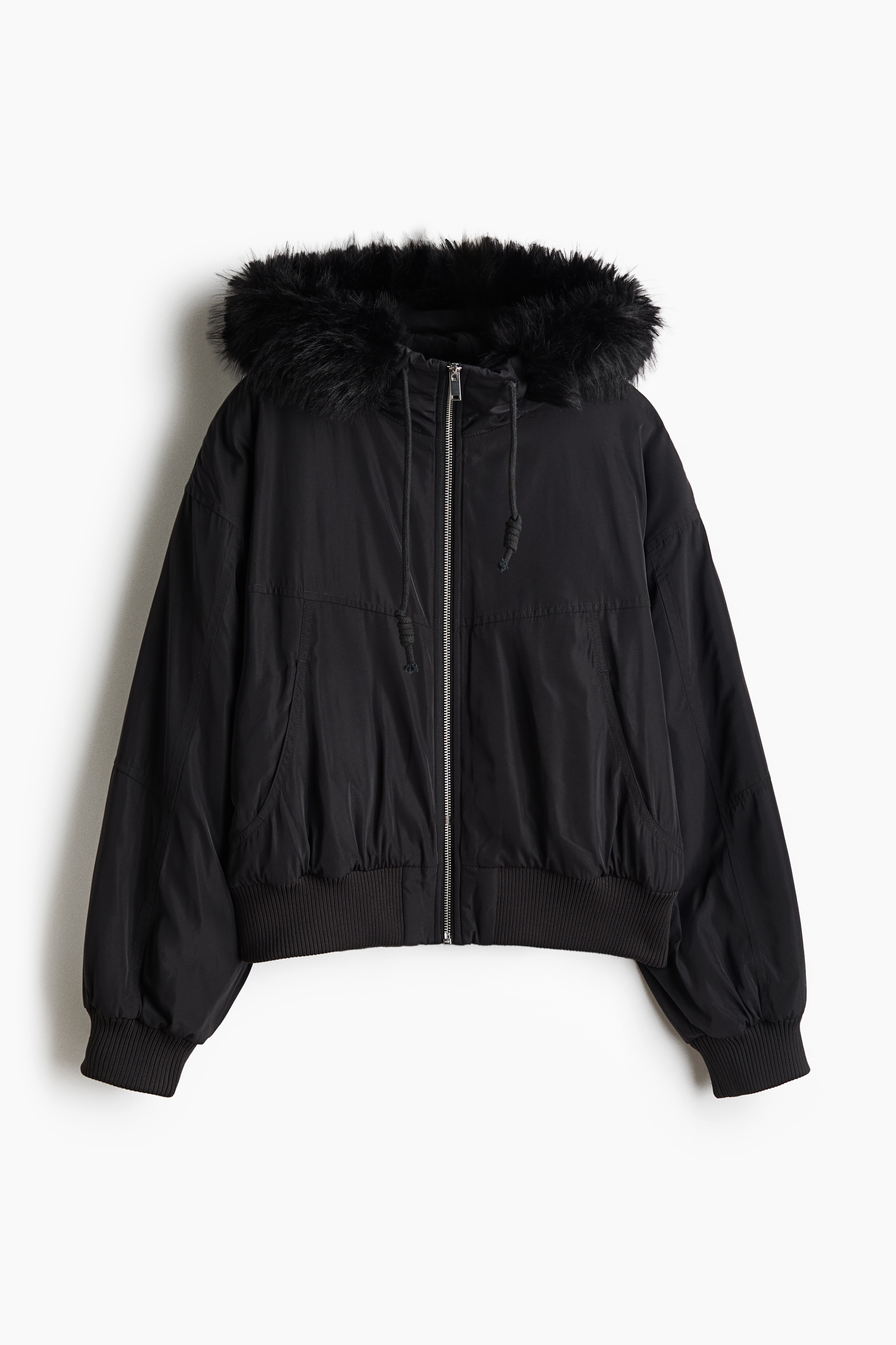 An oversized, dark-colored nylon jacket featuring a front zipper, ribbed hem, and a drawstring hood with fluffy trim is being worn by the model. The jacket is styled open over a cropped top and plaid trousers, with the hood pulled up over a baseball cap.