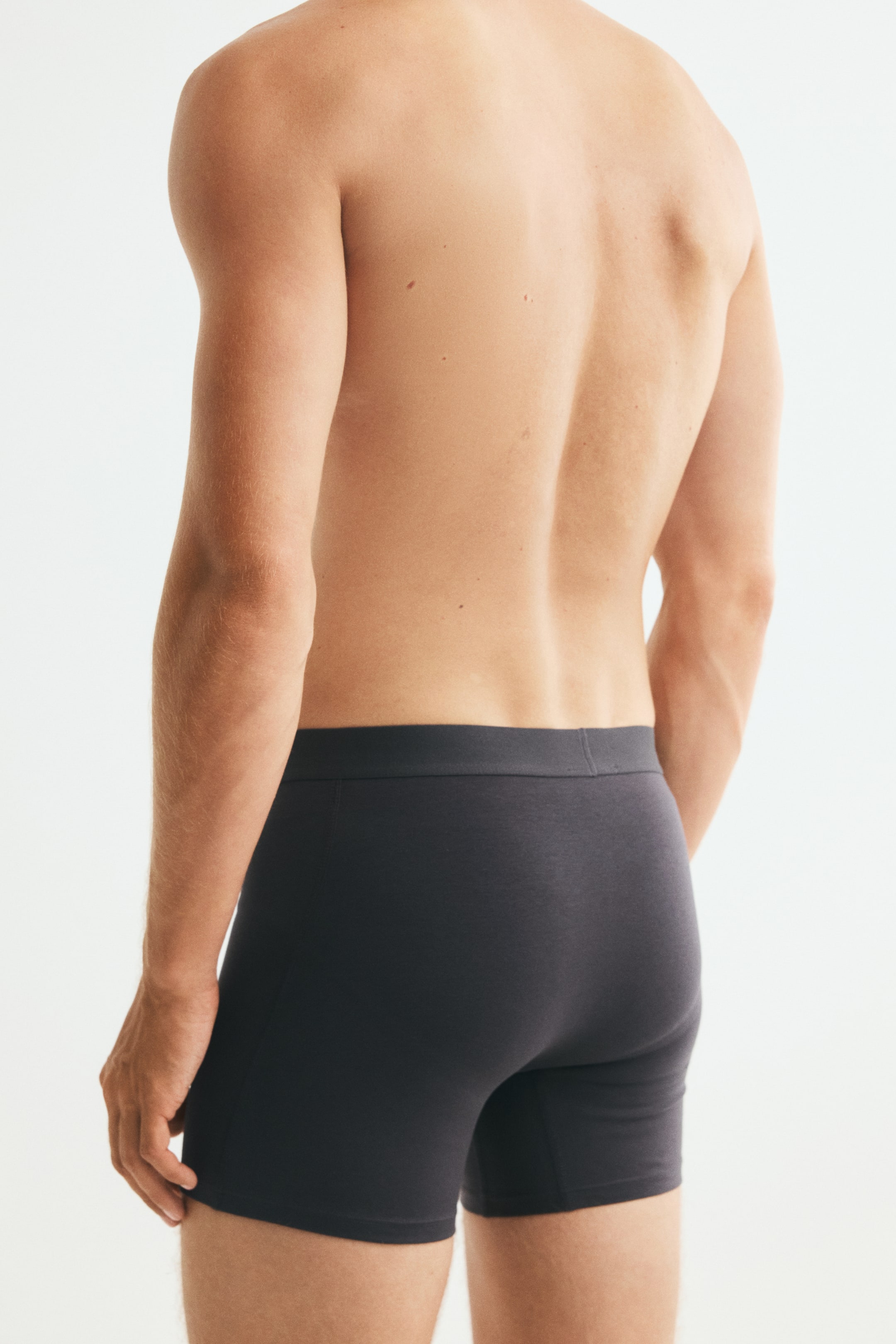 View larger image: Dark grey boxer briefs featuring a flat elastic waistband are being worn by the model. The close-fitting underwear extends to mid-thigh and is shown from a rear three-quarter angle.