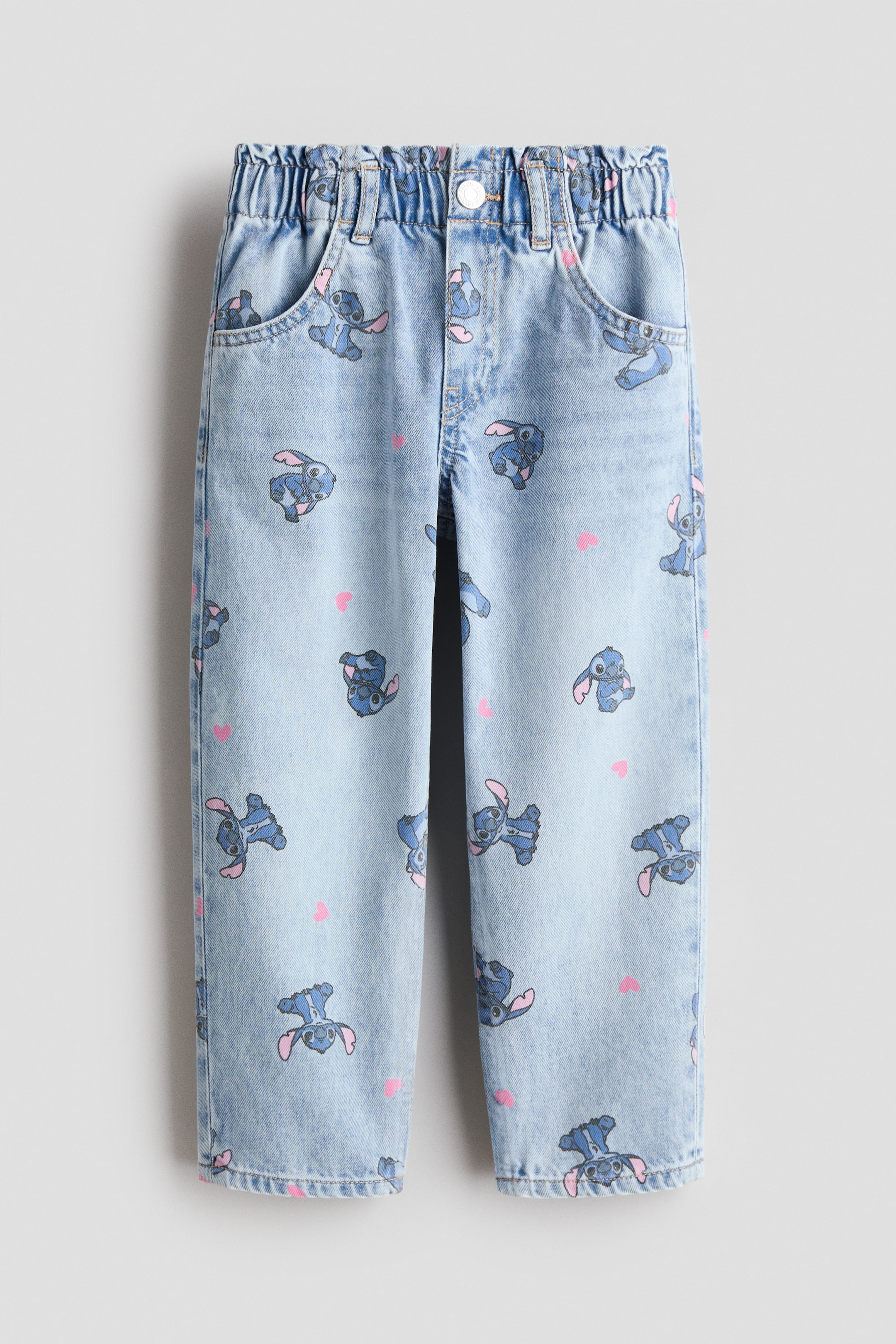 Ver imagen más grande: Pantalones vaqueros vistos de frente en suave denim de algodón que muestran un lavado azul claro con un estampado integral de personajes de dibujos animados azules y pequeños corazones rosas. Presentan un corte holgado en cadera y muslo con perneras cónicas, cintura paperbag con elástico ajustable, falsa bragueta, botón a presión y bolsillos delanteros abiertos.