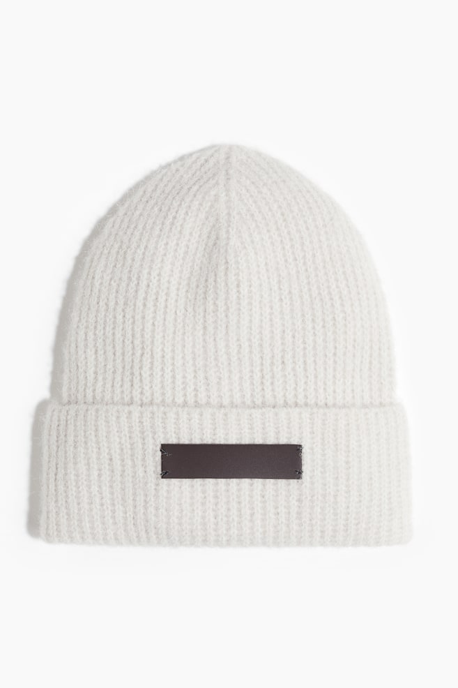 A white knit beanie is shown from the front, featuring a ribbed texture and a folded brim. A rectangular dark brown patch is sewn onto the center of the brim, with visible stitching at the corners. - 1