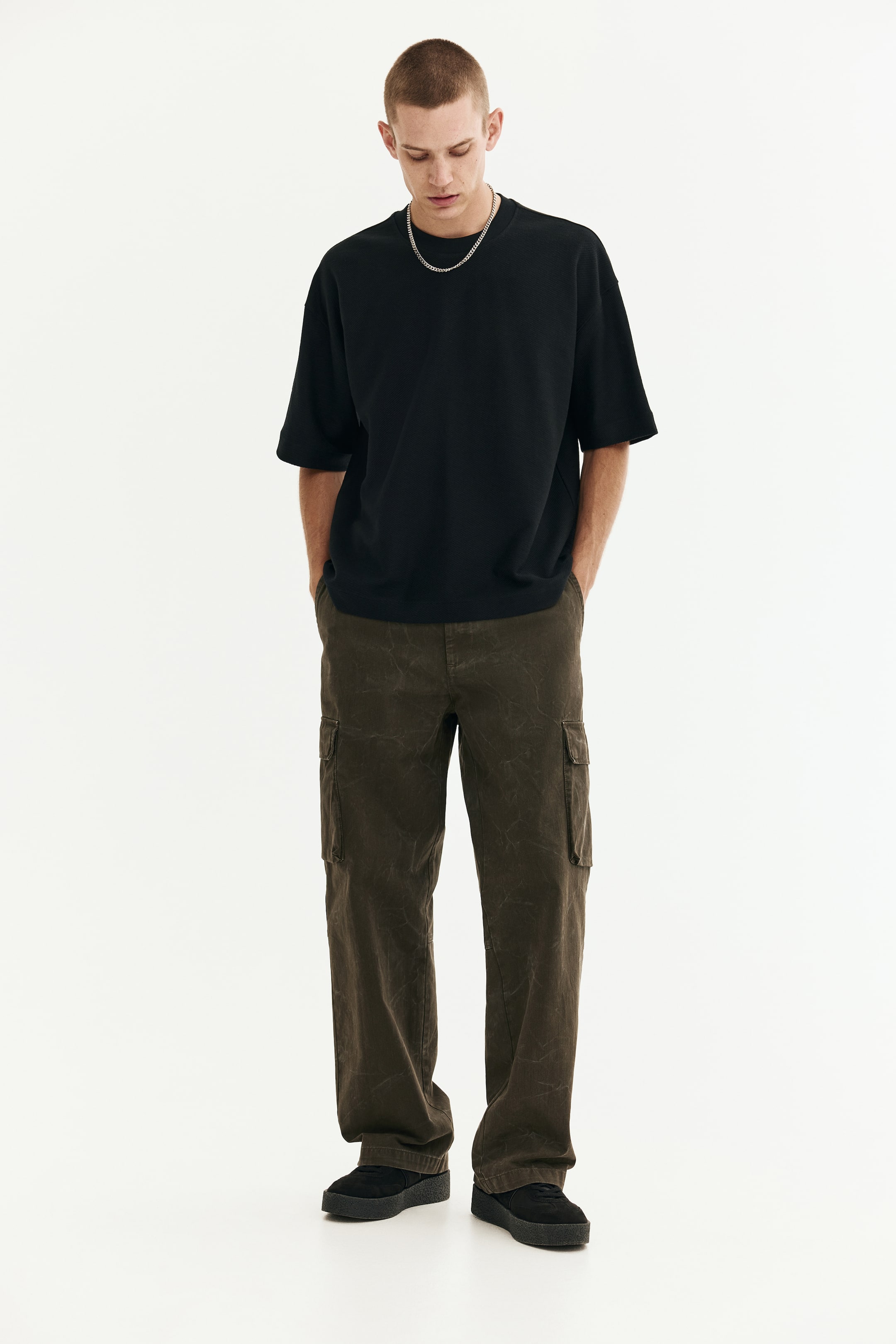 View larger image: A black oversized t-shirt is being presented from a front view, featuring a crew neck and short sleeves. It is being paired with dark brown relaxed-fit cargo pants, which have side utility pockets and a faded texture, along with black suede-like shoes with thick soles.