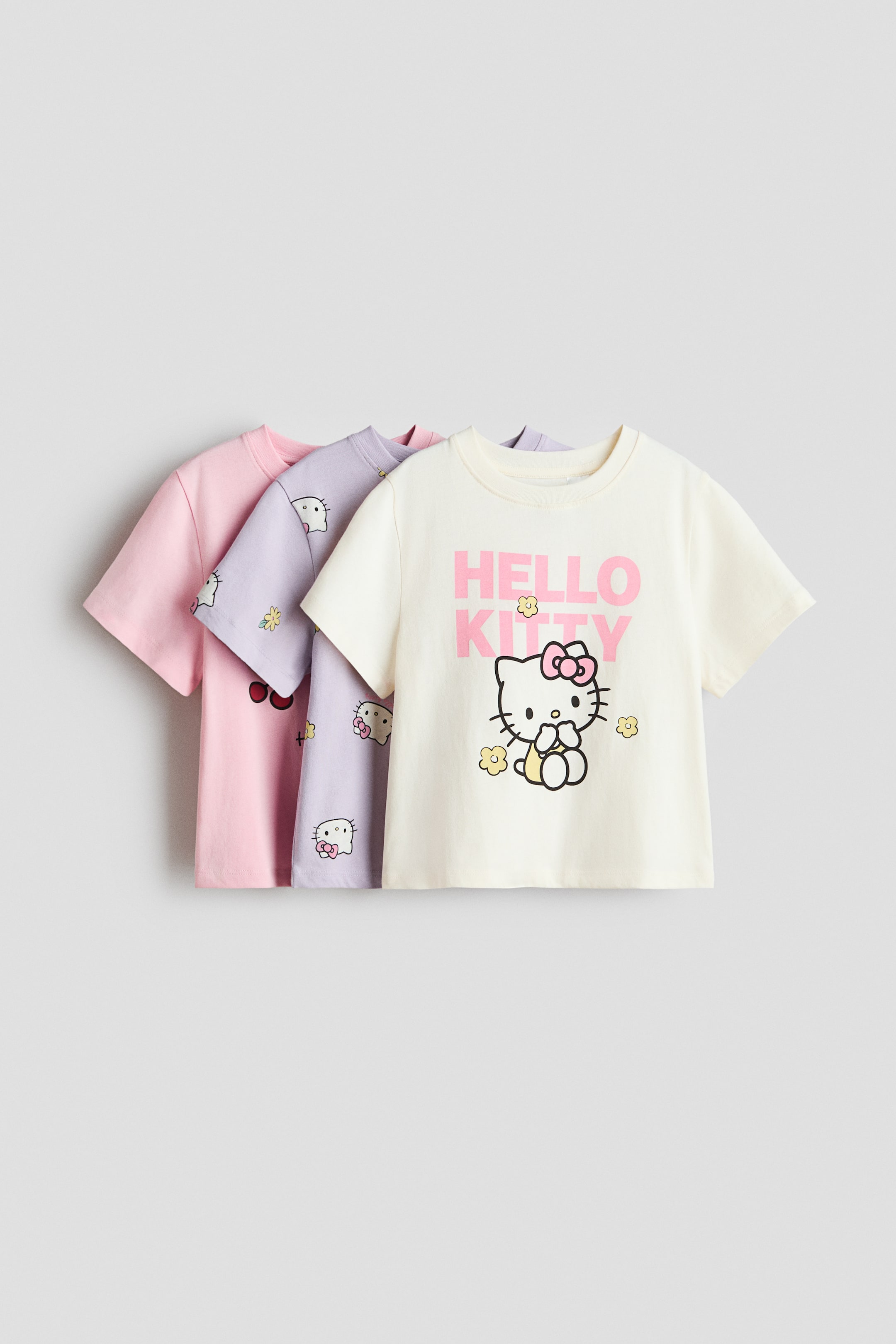 View larger image: Three short-sleeved t-shirts are showcased in a front-facing and layered arrangement, transitioning from cream to light purple to light pink. The cream t-shirt highlights a pink "HELLO KITTY" graphic featuring the character, while the light purple t-shirt presents an all-over print of Hello Kitty heads and flowers. The light pink t-shirt exhibits a cherry print.