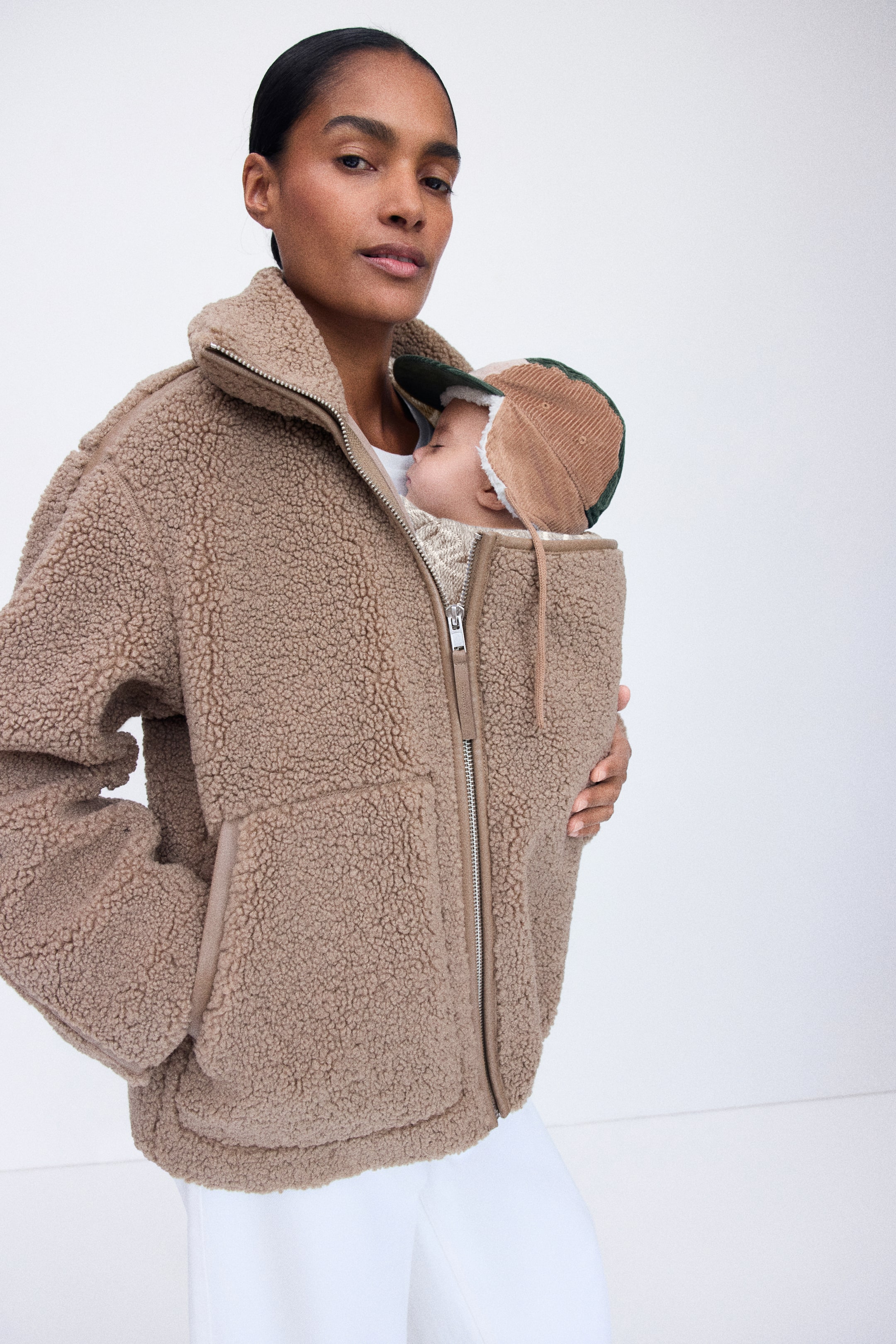 Se større billede: En oversized teddyjakke i lysebrun bæres af modellen og har en høj funnelkrave, store sidelommer samt en central lynlåslukning med integreret babybærepanel. Modellen bærer en baby, der er fastgjort inde i frontpanelet, mens hun støtter den med den ene hånd.
