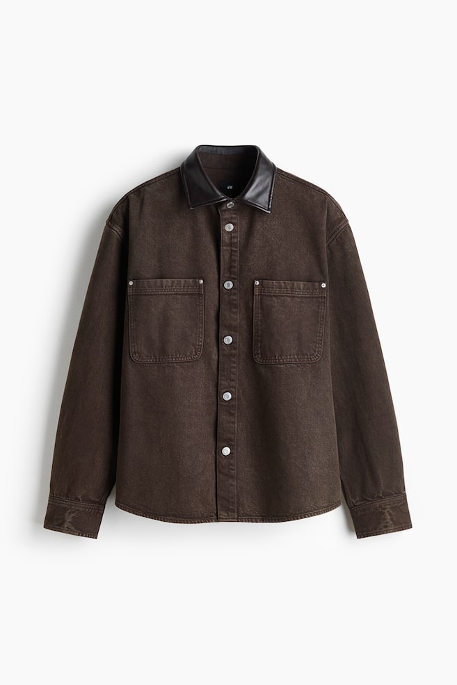En mörkbrun denim skjortjacka visas framifrån, med en kontrasterande krage i svart läderlook och en främre knäppning med silvriga knappar. Jackan har två stora bröstfickor utanpå med detaljer i silverfärgade nitar samt långa ärmar med manschetter och knappar. - 1