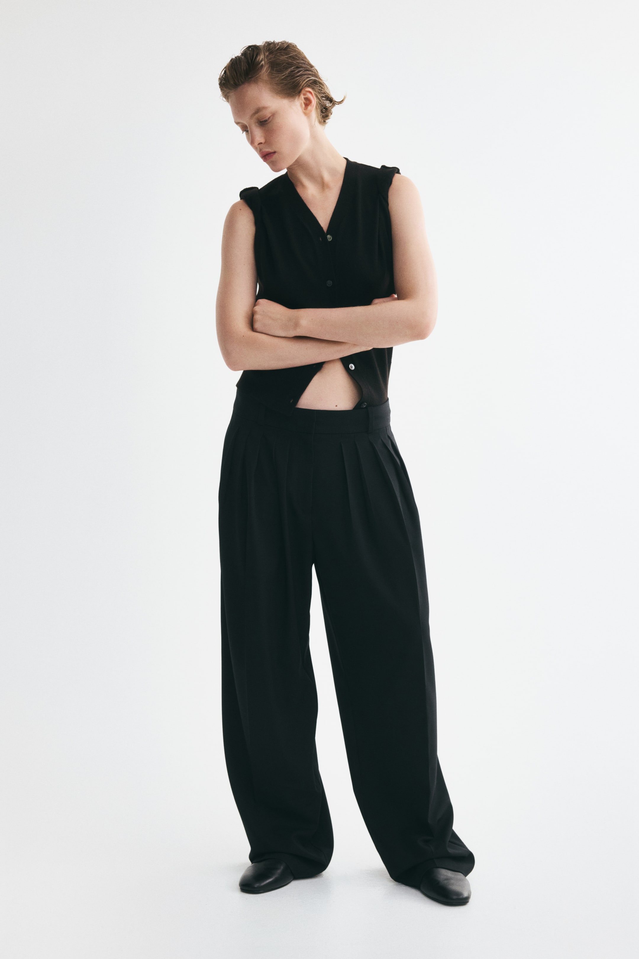 View larger image: Loose-fit black trousers in woven fabric, featuring a high waist, multiple front pleats, wide legs, and creases down the front, are being worn by the model, who is also wearing a black sleeveless top.