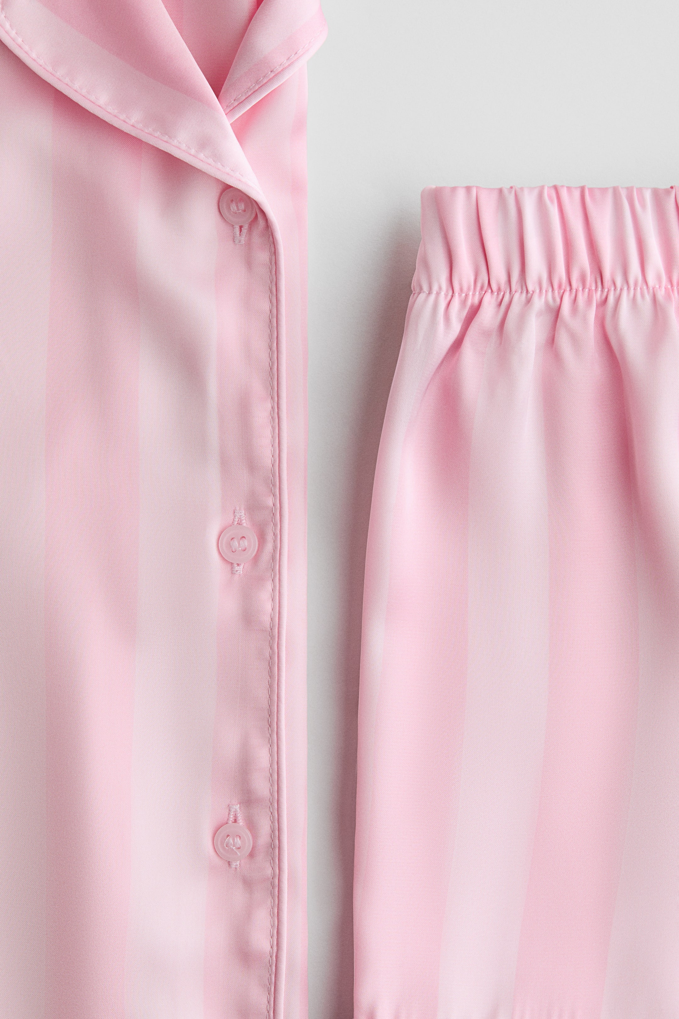 Näytä suurempi kuva: Pehmeästä vaaleanpunaisesta satiinista valmistettu pyjamasetti esitetään EDESTÄ, ja siinä on kauluksellinen paita ja nappilista, sekä shortsit, joissa on peitetty joustava vyötärönauha. Molemmissa osissa on kauttaaltaan pystysuuntainen raidoitus eri sävyissä vaaleanpunaista.