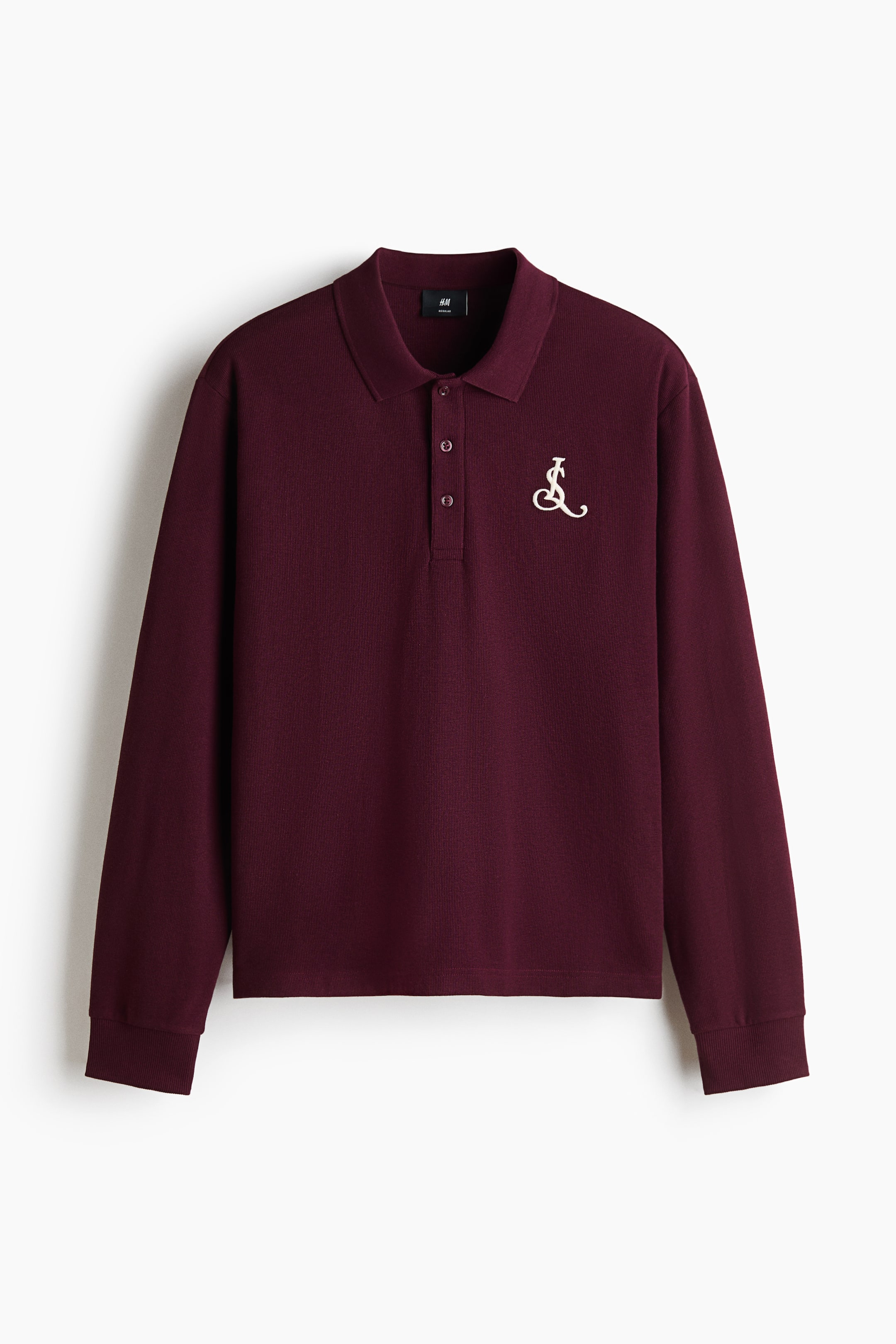 View larger image: A long-sleeved burgundy polo shirt in a textured waffle knit is shown from the front. It features a ribbed collar, a three-button placket, ribbed cuffs, and a small cream-coloured embroidered monogram on the left chest.
