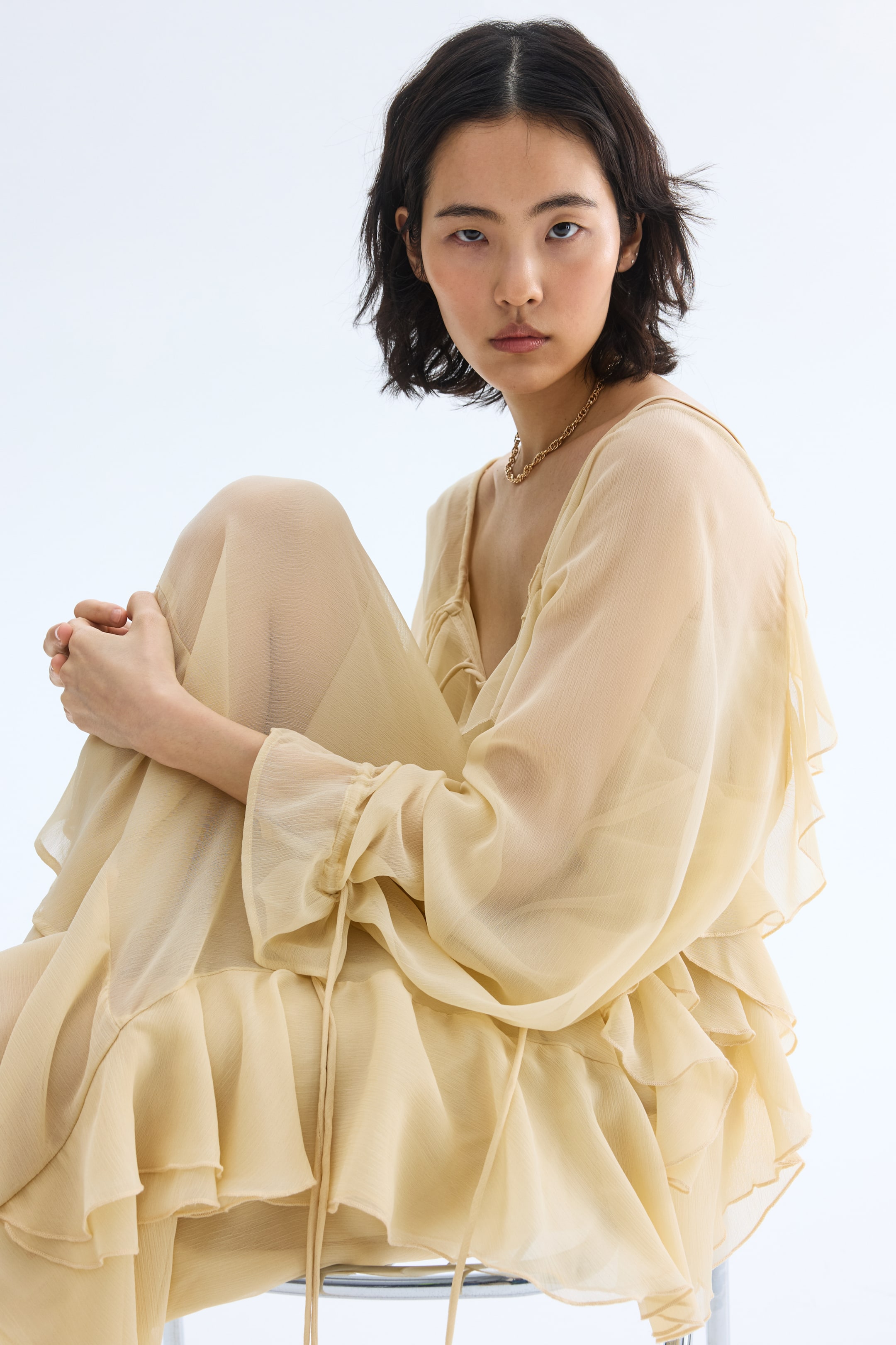 View larger image: A light beige sheer dress featuring a v-neckline and long, full sleeves with gathered cuffs. The dress is detailed with multiple tiers of flowing ruffles throughout the sleeves and skirt, with visible ties gathering the fabric at the side.