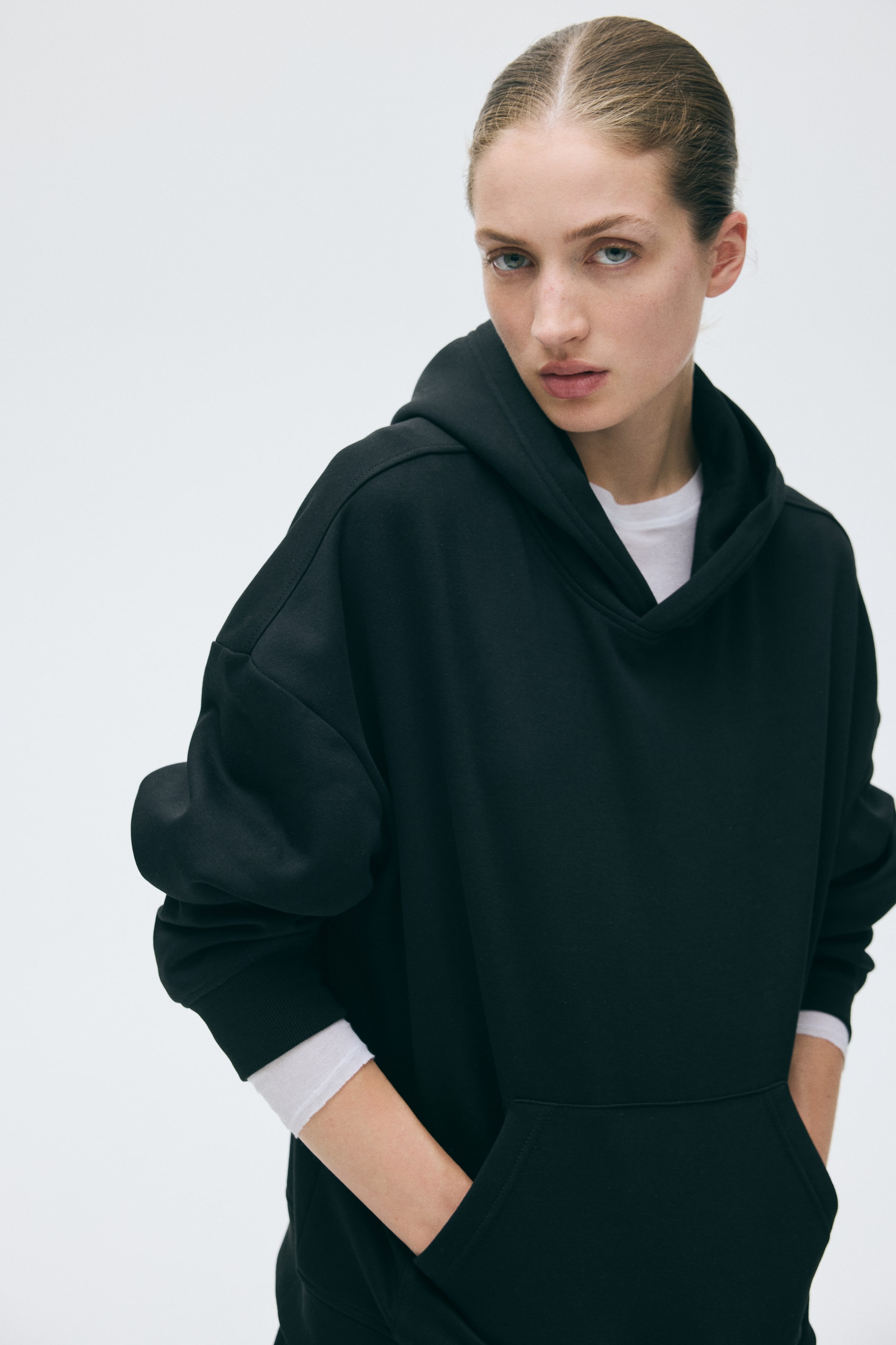 Grotere afbeelding bekijken: Een zwarte hoodie met een dubbele laag capuchon, laag hangende schouders en geribde boorden wordt gedragen door het model. De handen van het model zijn in de kangoeroezak geplaatst, en een wit topje is zichtbaar als laag eronder bij de halslijn en boorden.