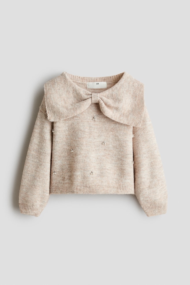 Se muestra de frente un jersey de punto melange beige claro, con un gran cuello decorativo con lazo y detalles de perlas sintÃ©ticas dispersas en el cuerpo. El diseÃ±o incluye mangas largas con puÃ±os de canalÃ© y bajo de canalÃ© a juego. - 1