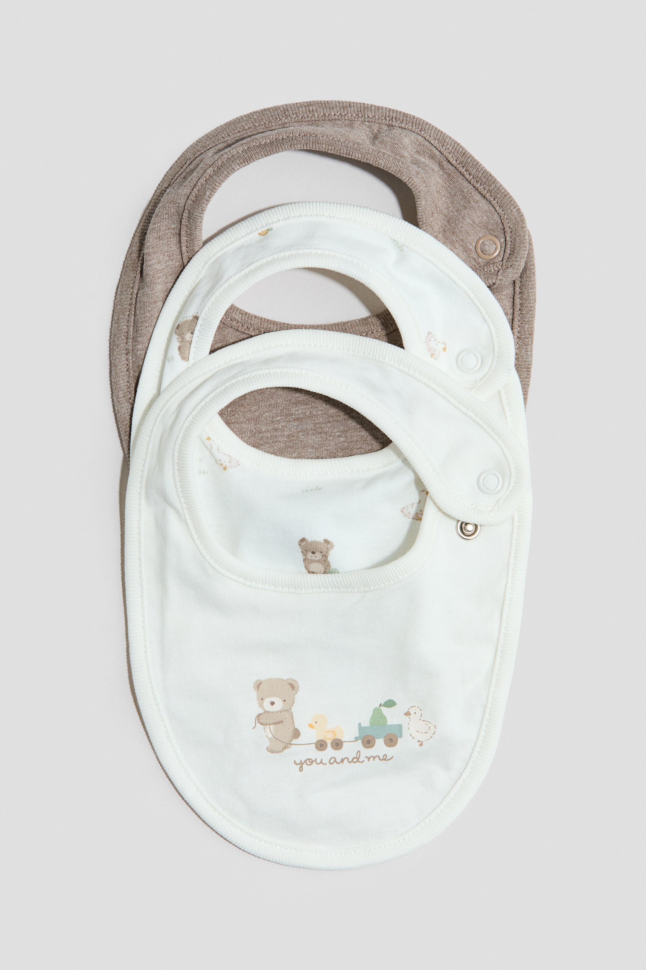 View larger image: Bibs crafted from soft cotton jersey, featuring an adjustable press-stud fastening on one side, are showcased from the front. The top off-white bib displays a charming pattern of a teddy bear pulling a train with a duck and a pear, accompanied by the text 'you and me', while beneath, a plain brown bib is visible.
