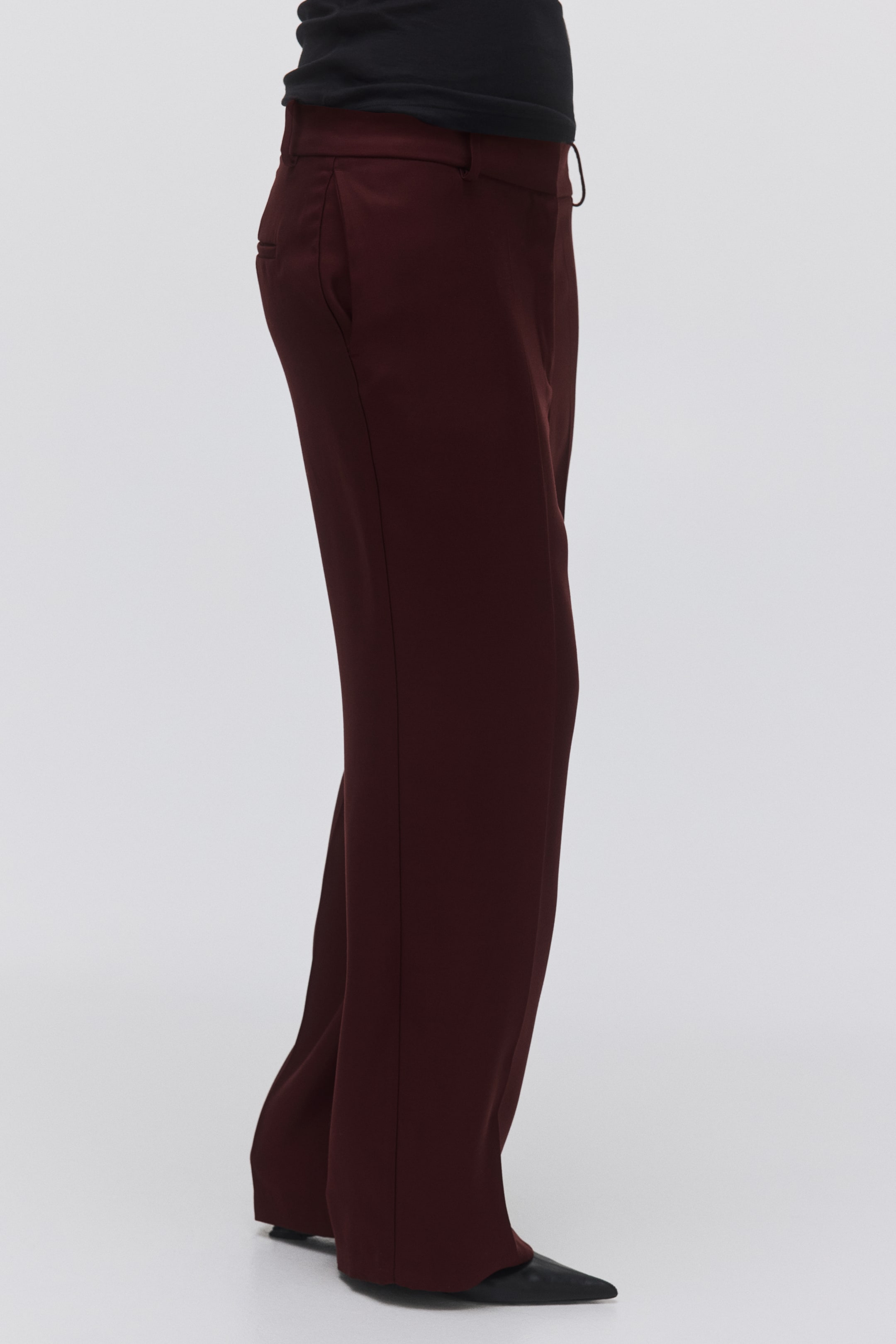 Agrandir l'image: Un pantalon large couleur marron est vu de côté, comportant une ceinture plate et une ouverture subtile de poche latérale au niveau de la hanche. Le tissu lisse tombe sur des chaussures pointues noires, avec un haut noir visible à la taille.