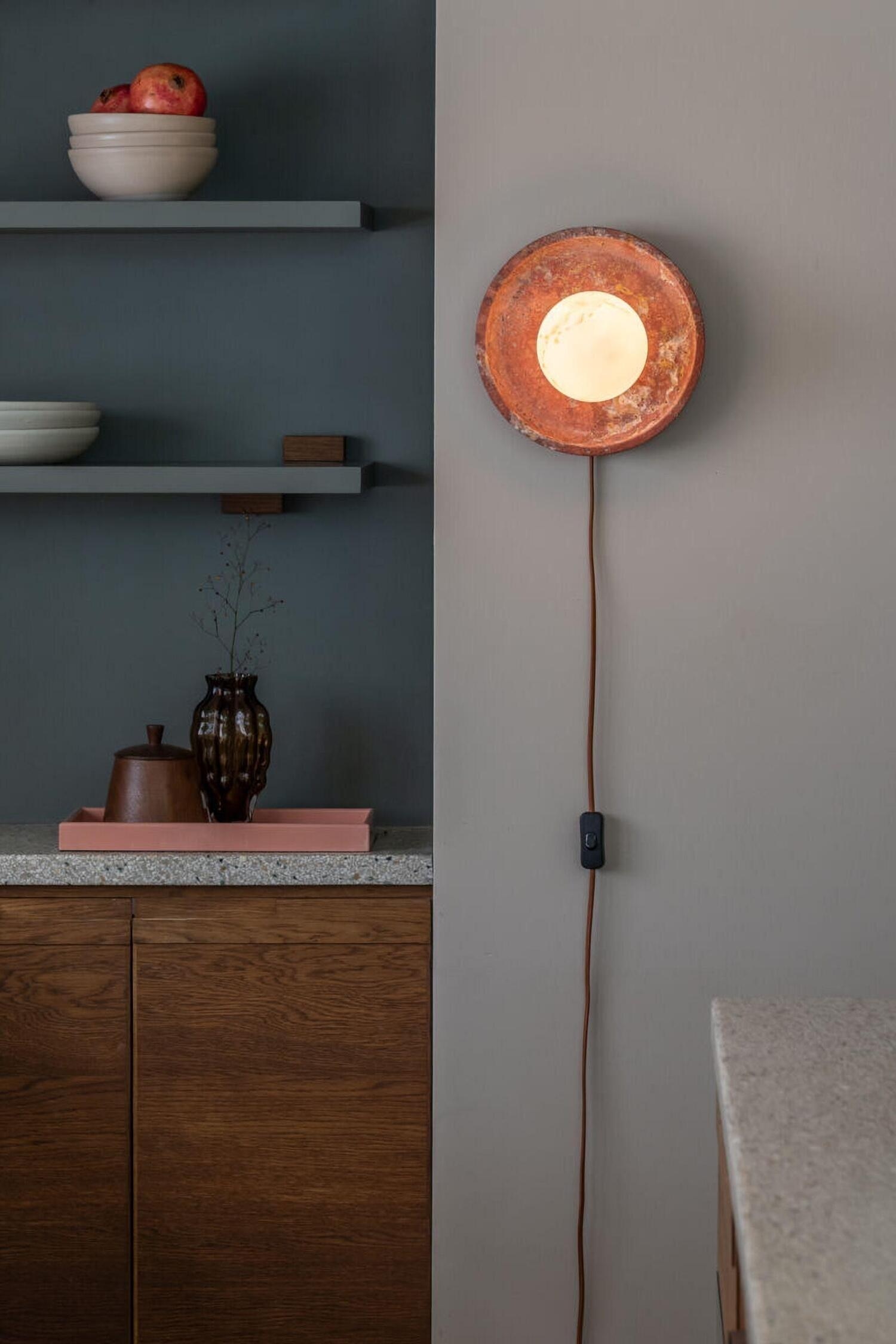 Een wandlamp wordt getoond vanuit een driekwart zijaanzicht, met een gloeiende, bolvormige glazen globe met subtiele gele marmering, gemonteerd op een ronde, gestructureerde roodbruine stenen achterplaat. Een bruine cilindervormige wandbevestiging draagt de armatuur, met een bijpassend stoffen snoer dat naar beneden loopt.